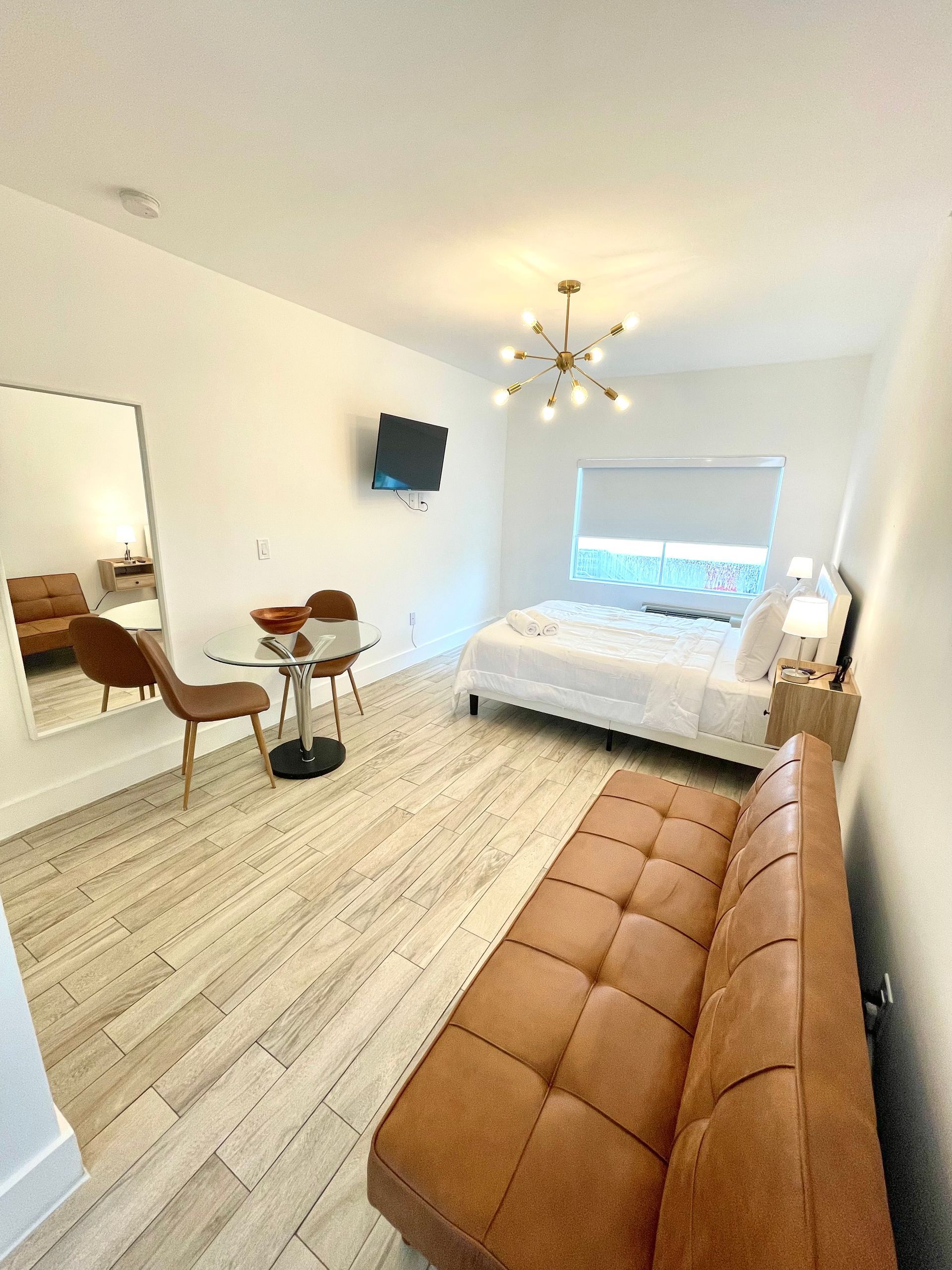 Small studio apartment with a bed, sofa, table, and mirror. Light wood flooring.