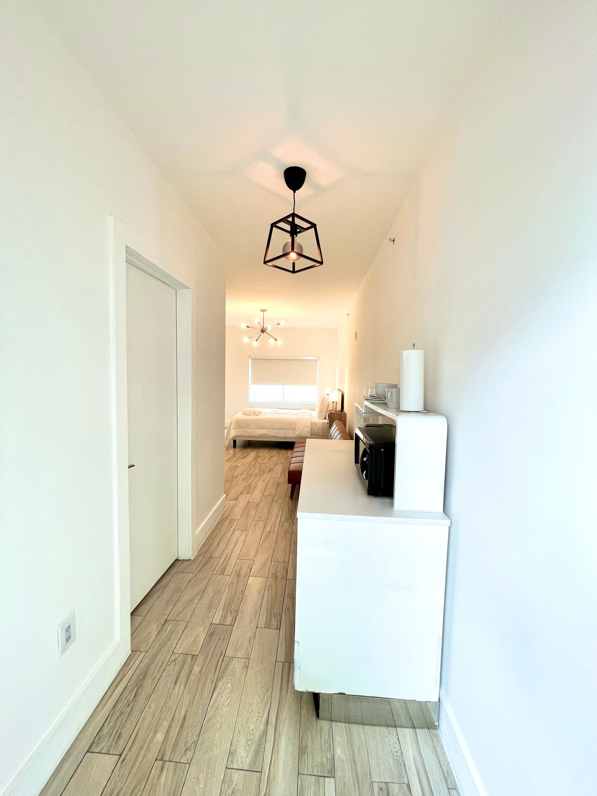 Narrow hallway with light wood floor, white walls, and a black pendant light.