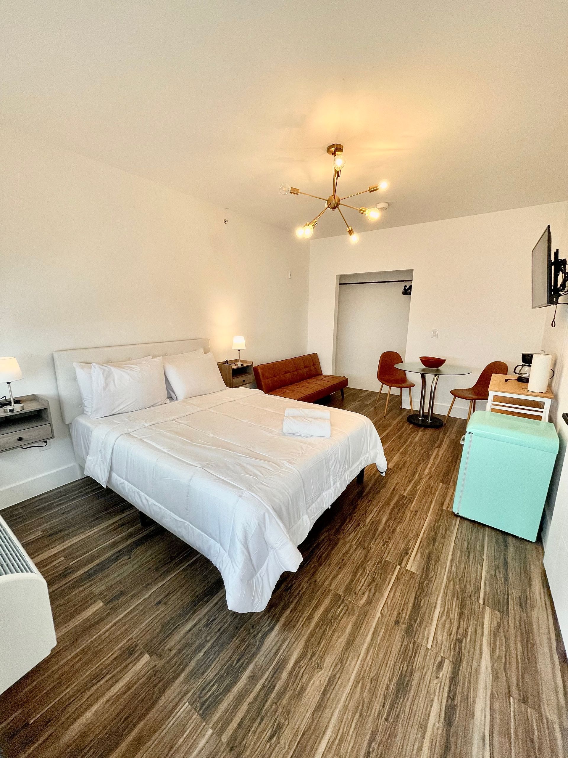 Bright, white hotel room with bed, seating area, and teal mini-fridge on wood-look floor.