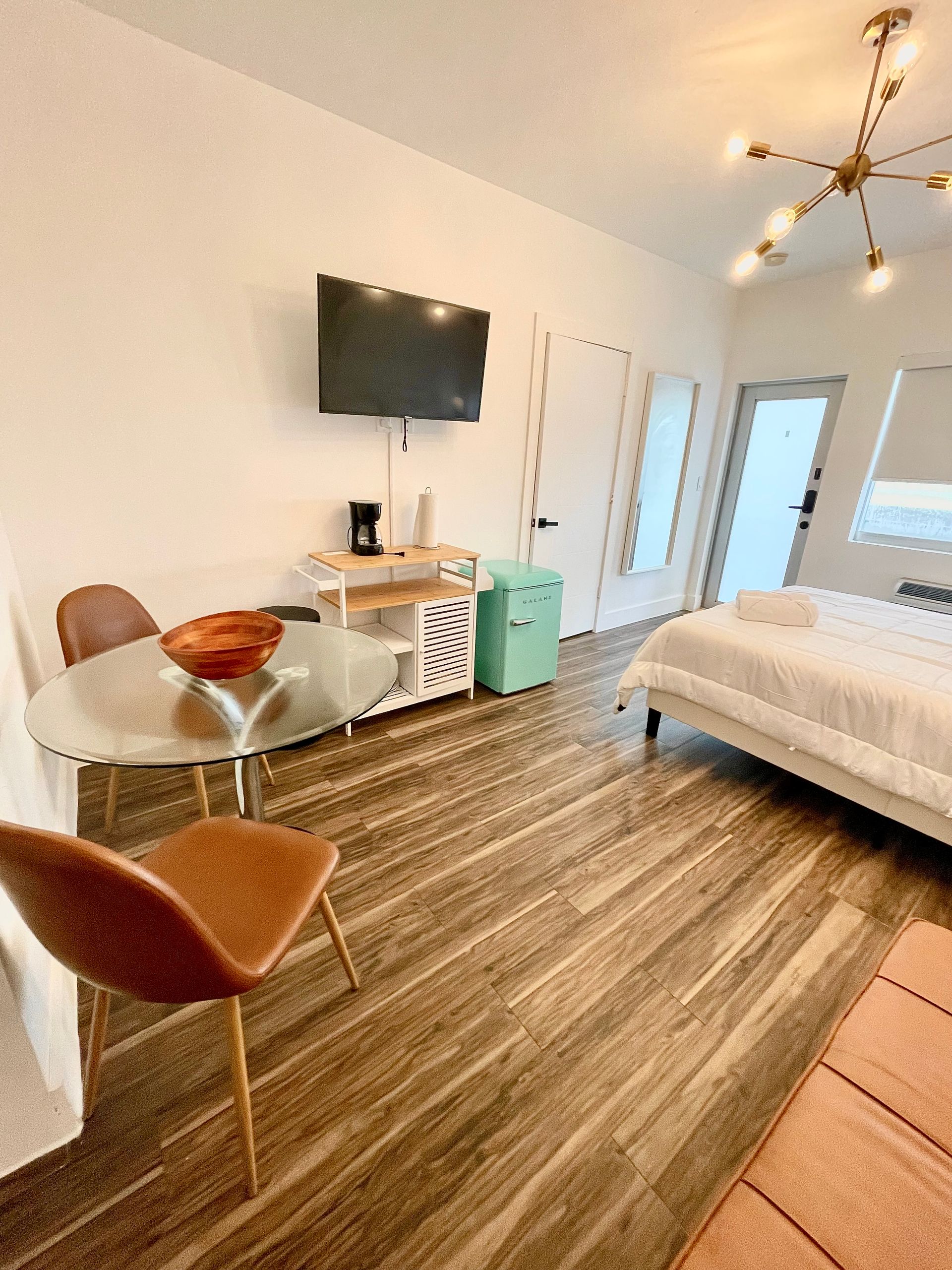 Bright studio apartment with wood floors, table, chairs, bed, TV, and small refrigerator.