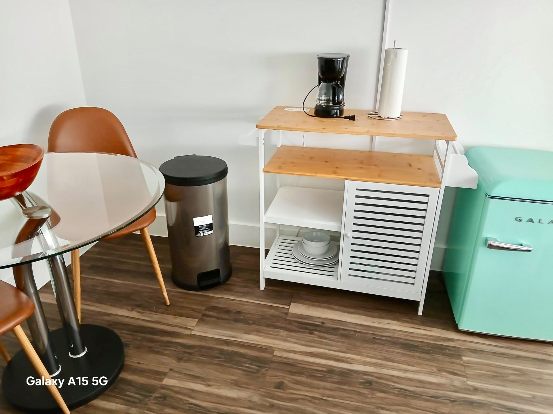 Small kitchen with round table, chair, counter with coffee maker, trash can, and turquoise refrigerator.