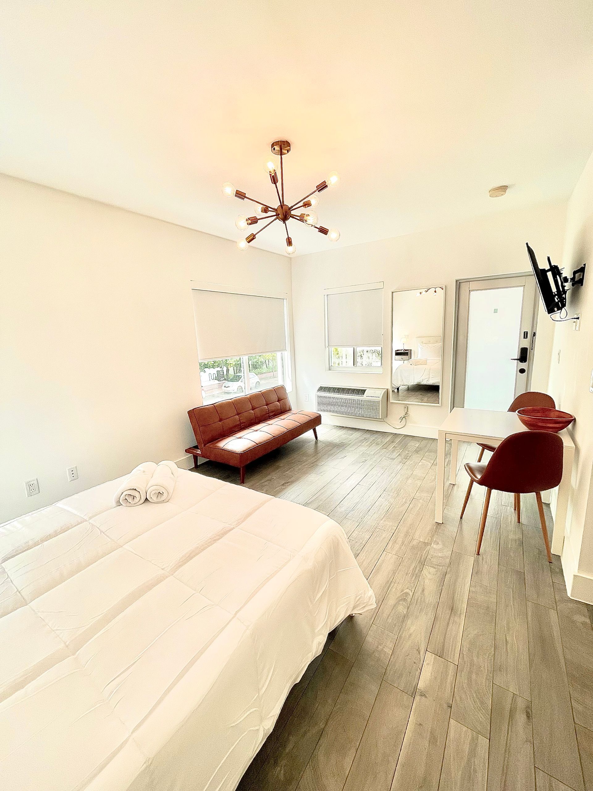 Bright studio apartment with a bed, red futon, dining set, and a chandelier.
