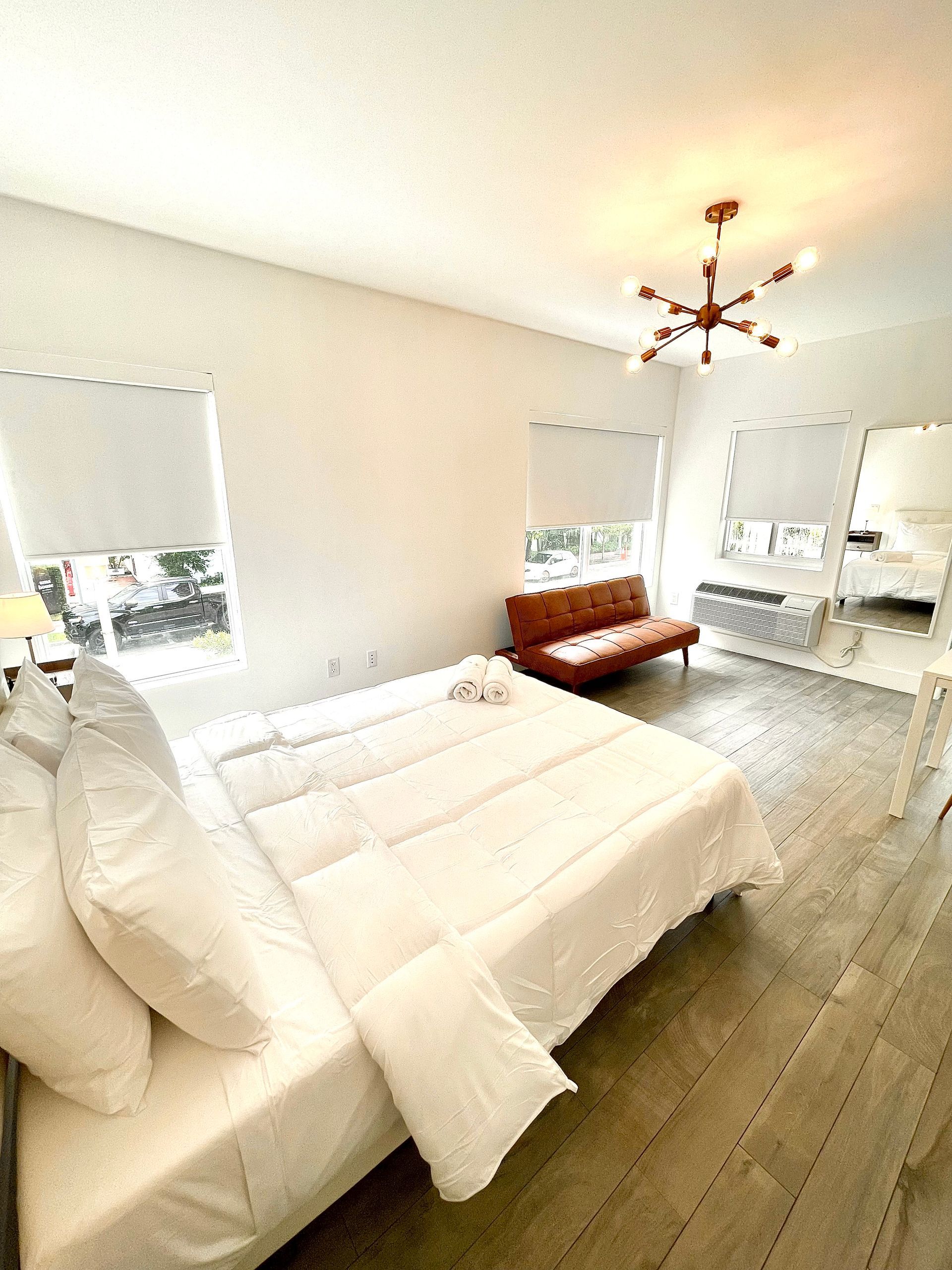 Bright bedroom with a white bed, brown sofa, windows, and a modern chandelier.