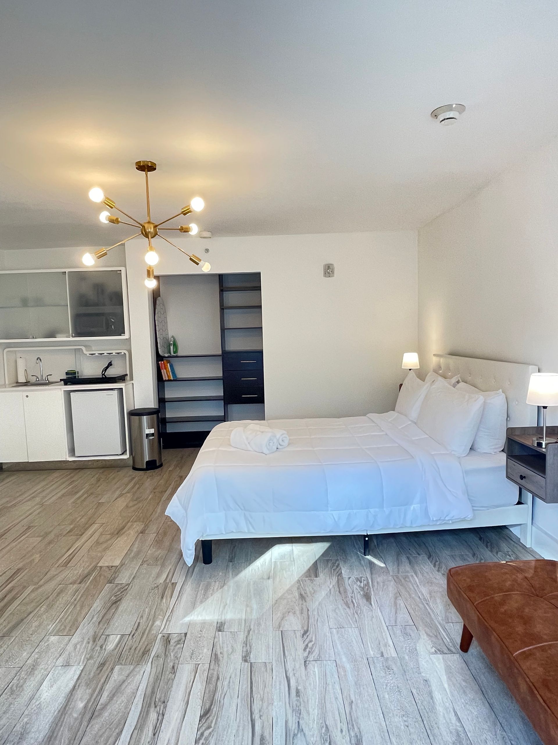 Bright studio apartment with a bed, small kitchen, and storage shelves; white and gold accents.