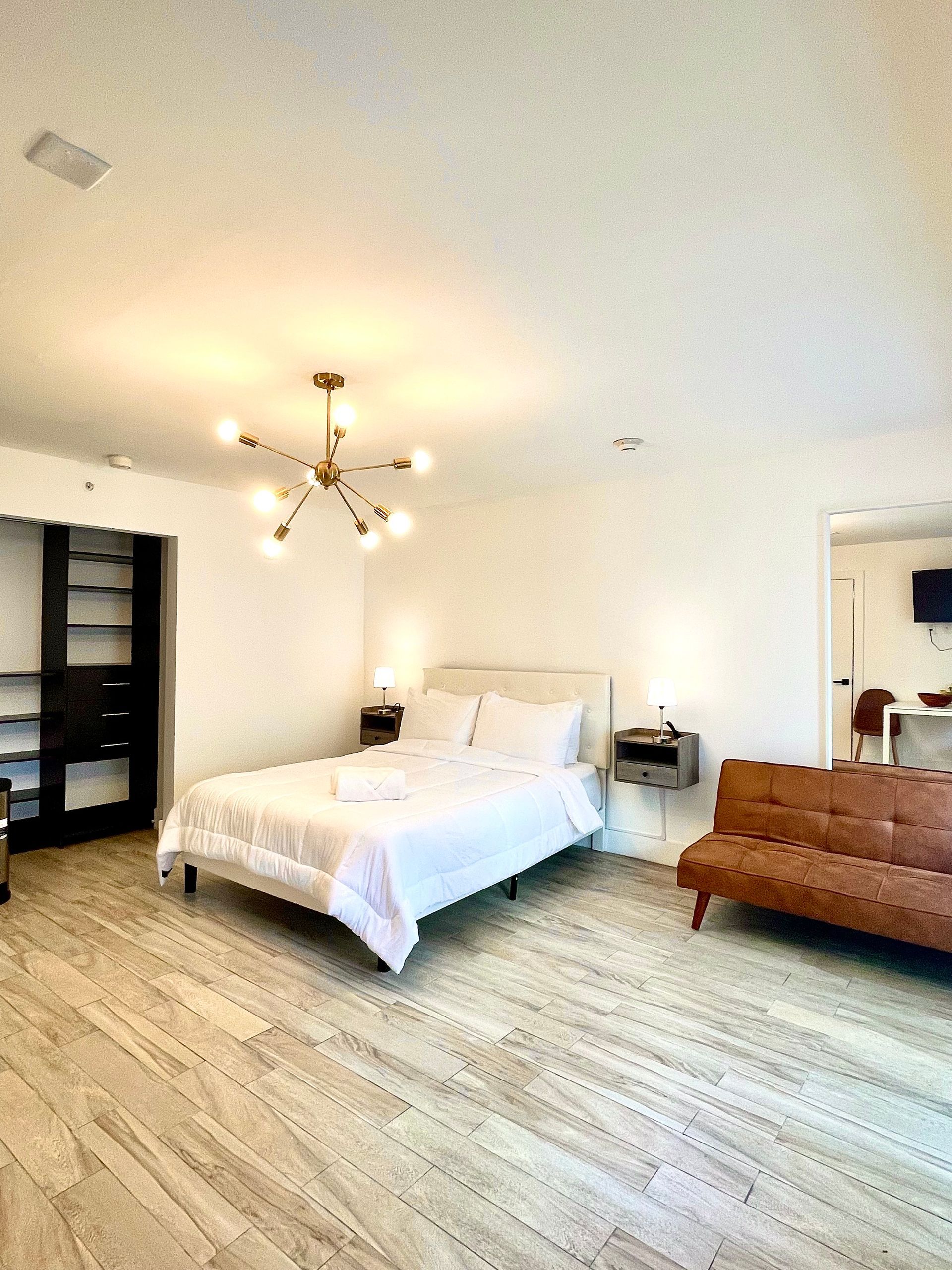 Bright bedroom with white walls, bed, modern lighting, brown sofa, and wood-like flooring.