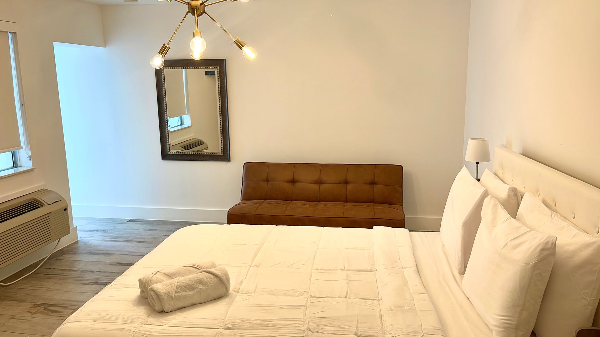 Bedroom with a bed, brown futon, mirror, and chandelier. White walls and bedding.