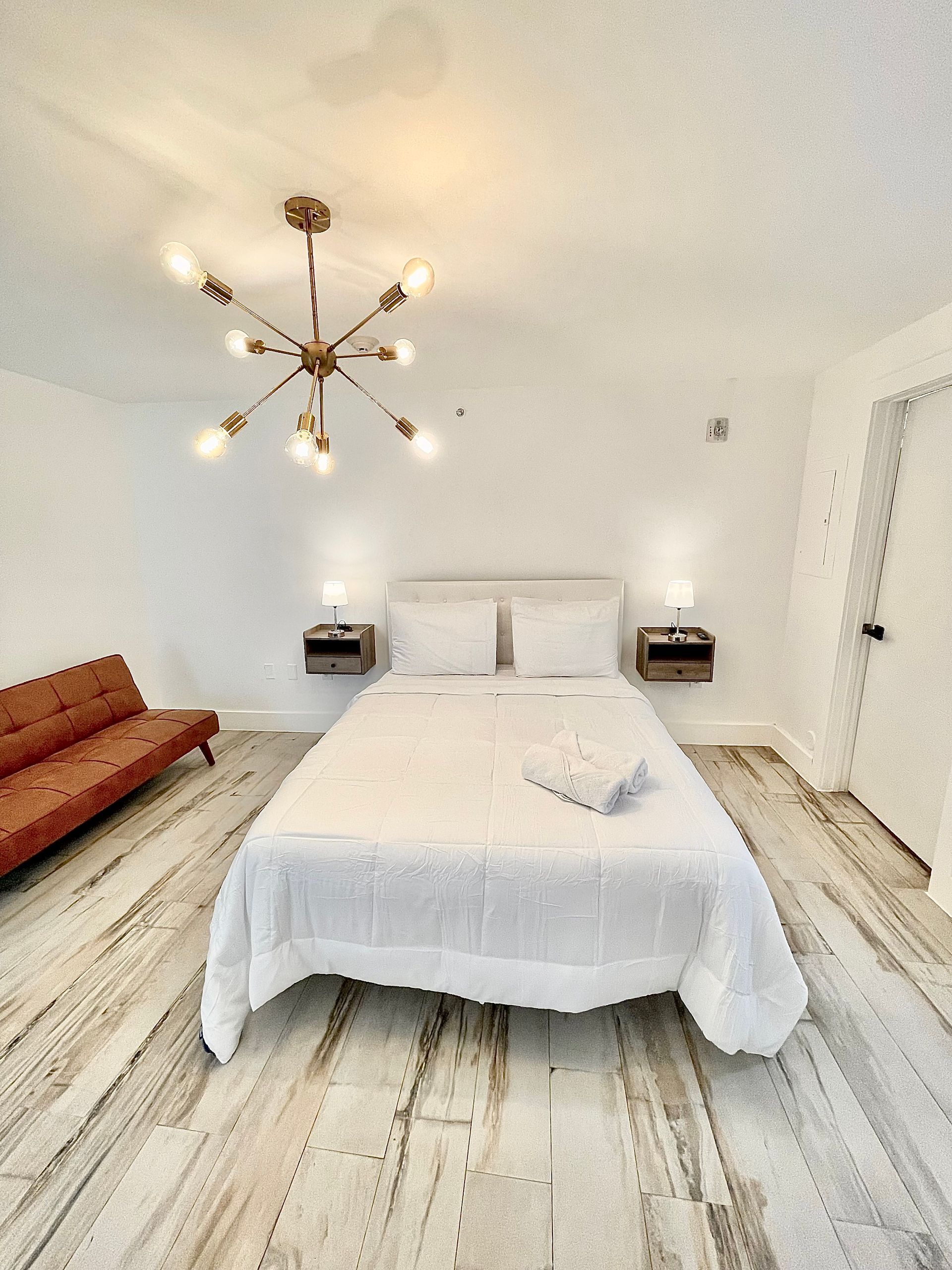 White bedroom with a bed, sofa, and gold chandelier. White walls, wood floor.