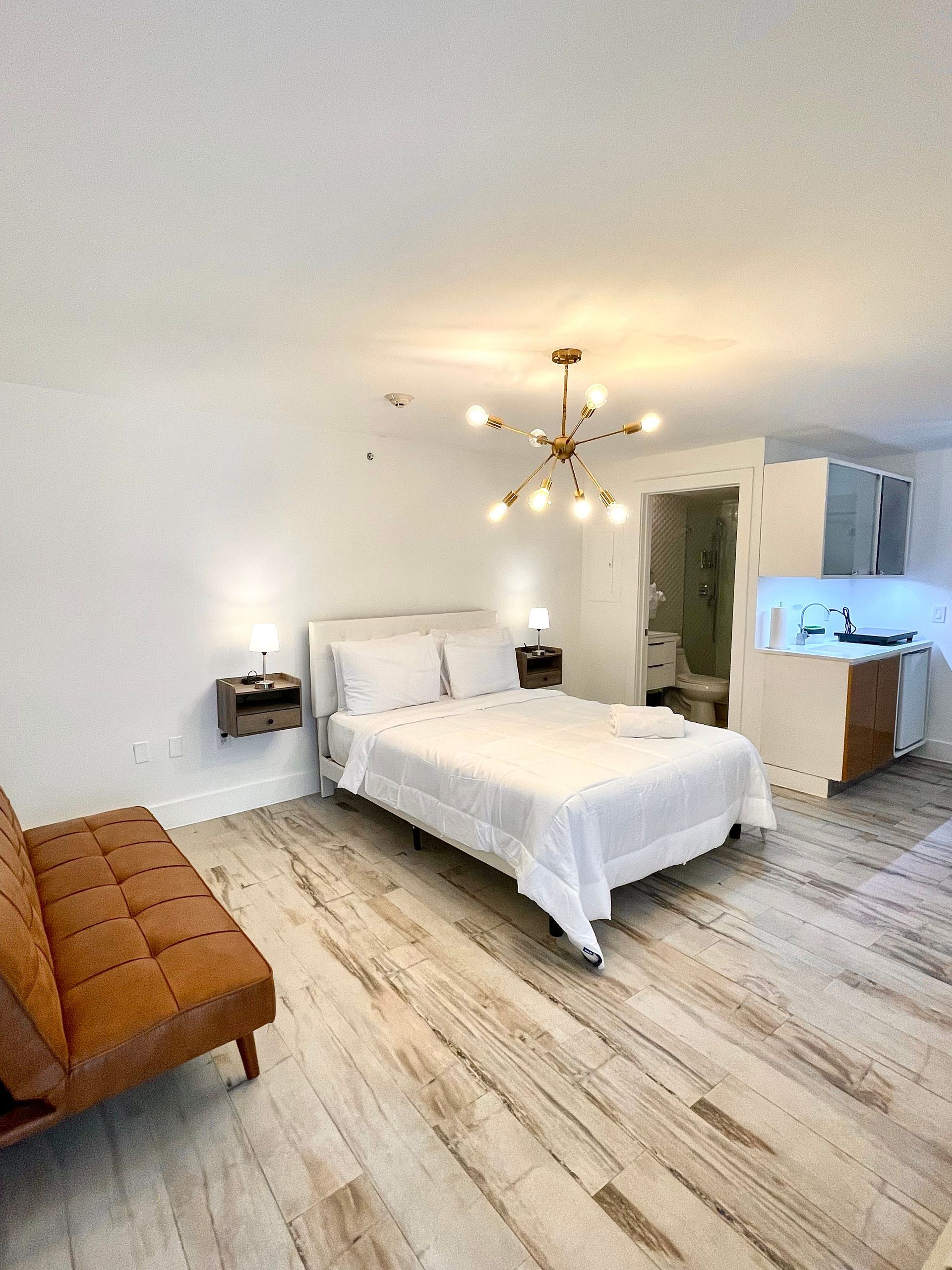 A studio apartment with a bed, sofa, kitchenette, and bathroom; white walls, wood floors.