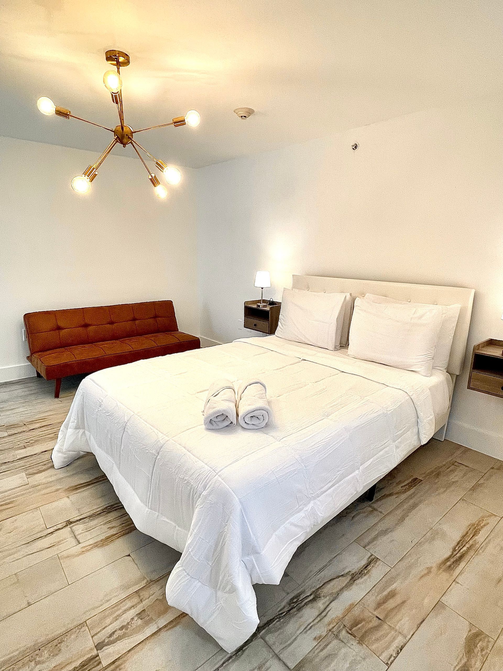 Bedroom with a bed, sofa, and modern light fixture. The room is brightly lit with white walls and a wooden floor.