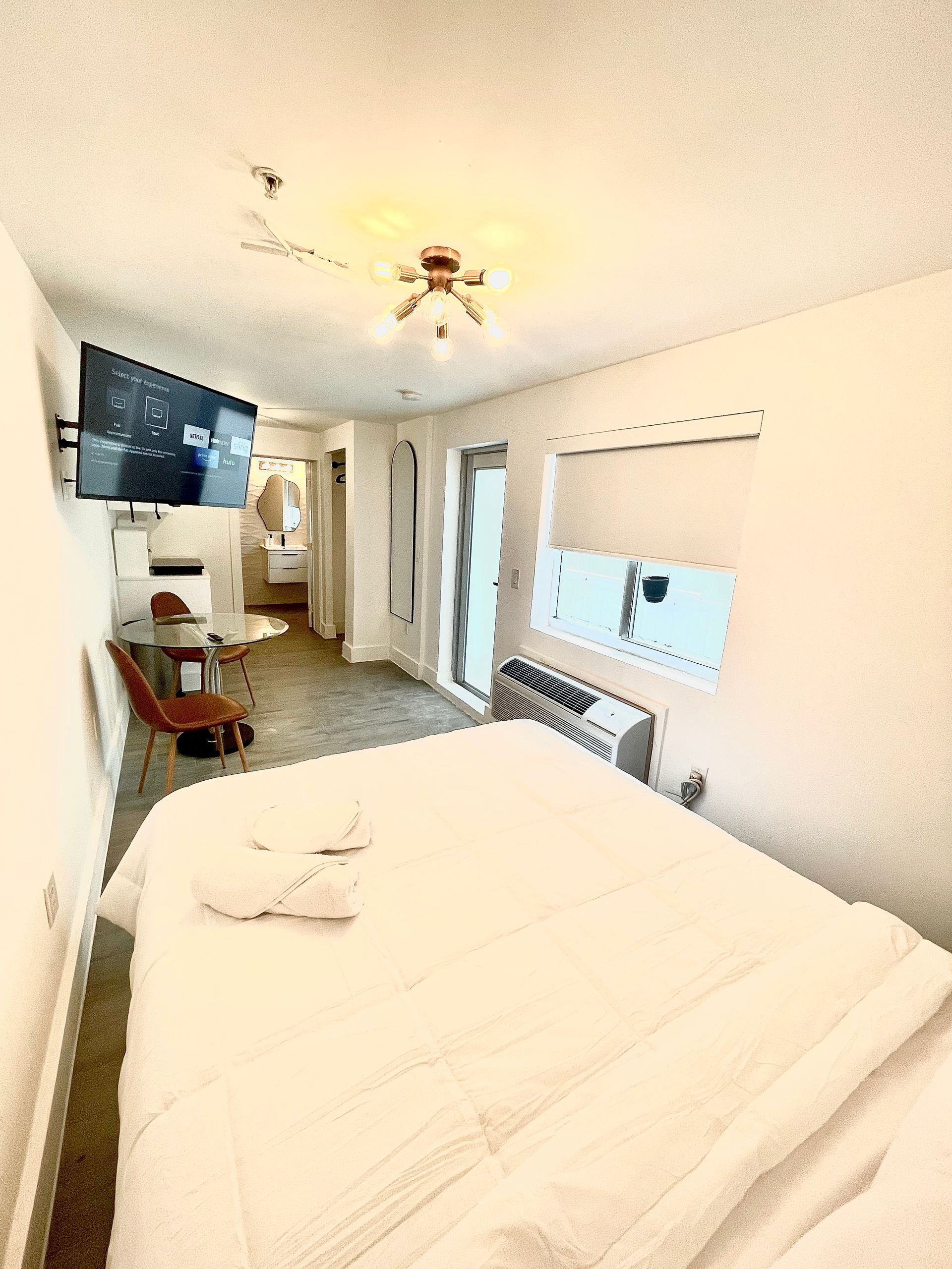 Bedroom with a bed, TV, and doorway to a bathroom. White walls, sunlight, and simple decor.