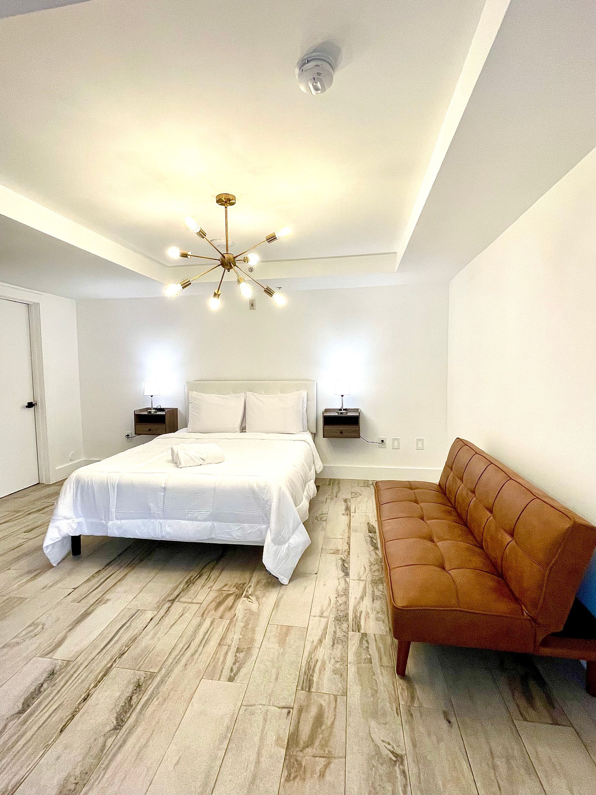 Bedroom with a bed, a brown sofa, and a gold chandelier. Light wood floors and white walls.