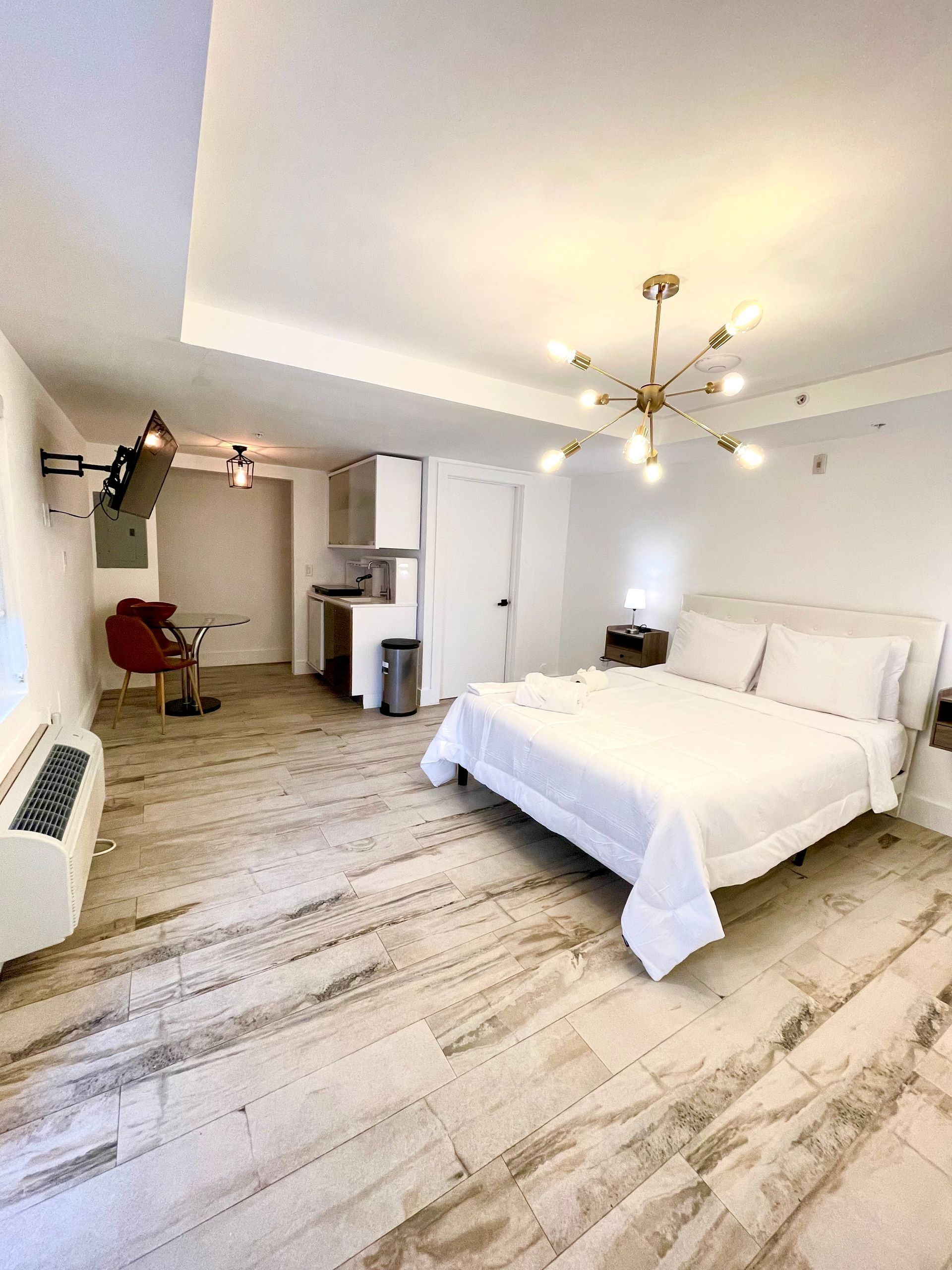 Bedroom with bed, kitchenette, and modern chandelier. White and beige tones with wood-like flooring.