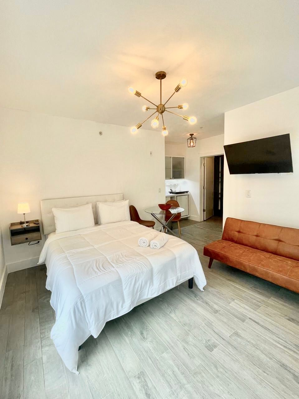 Bedroom with a bed, couch, TV, and kitchen area. White walls, gray floors, and a modern chandelier.