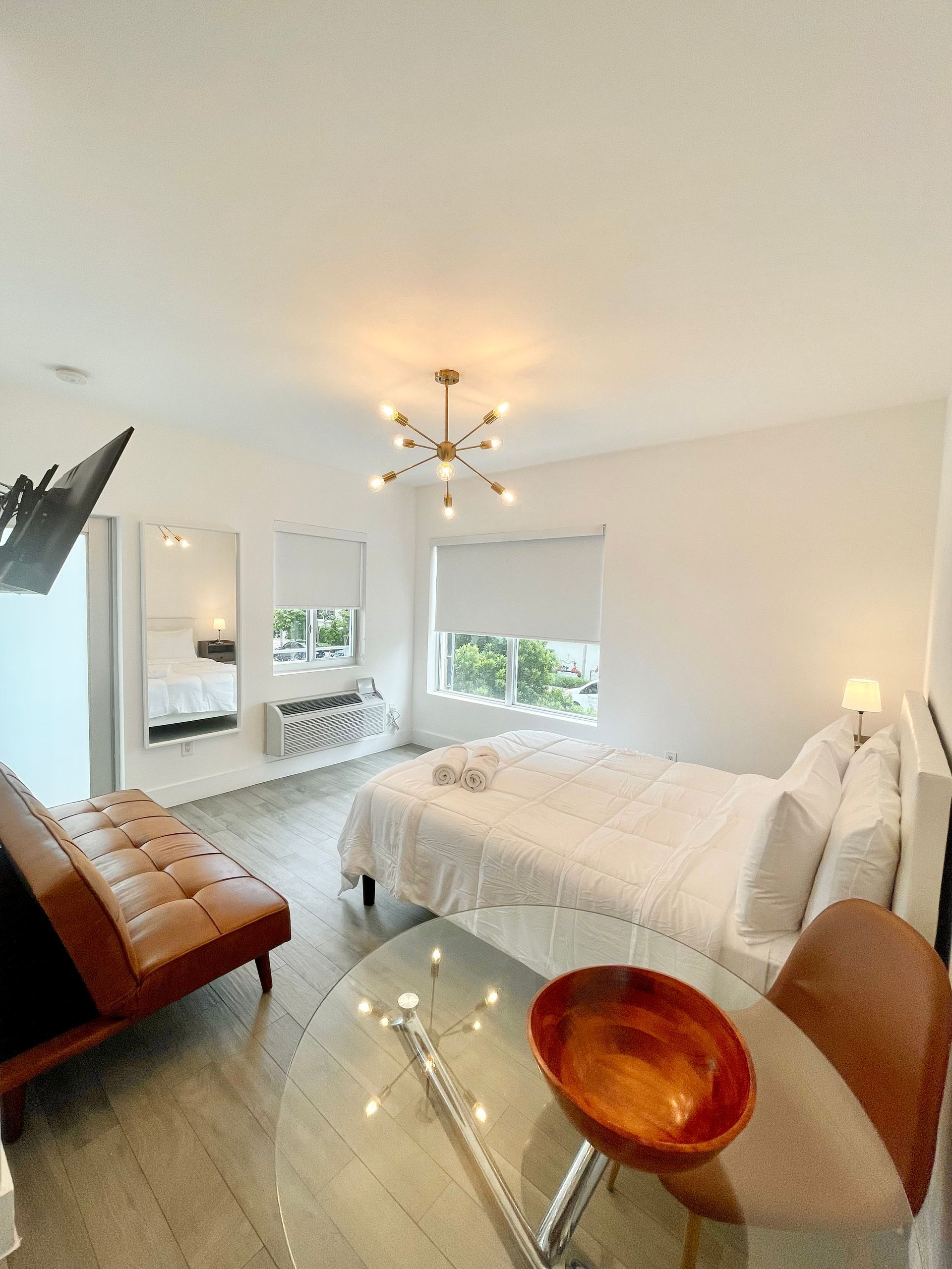 A modern studio apartment with a bed, sofa, and round glass table. White walls and decor.