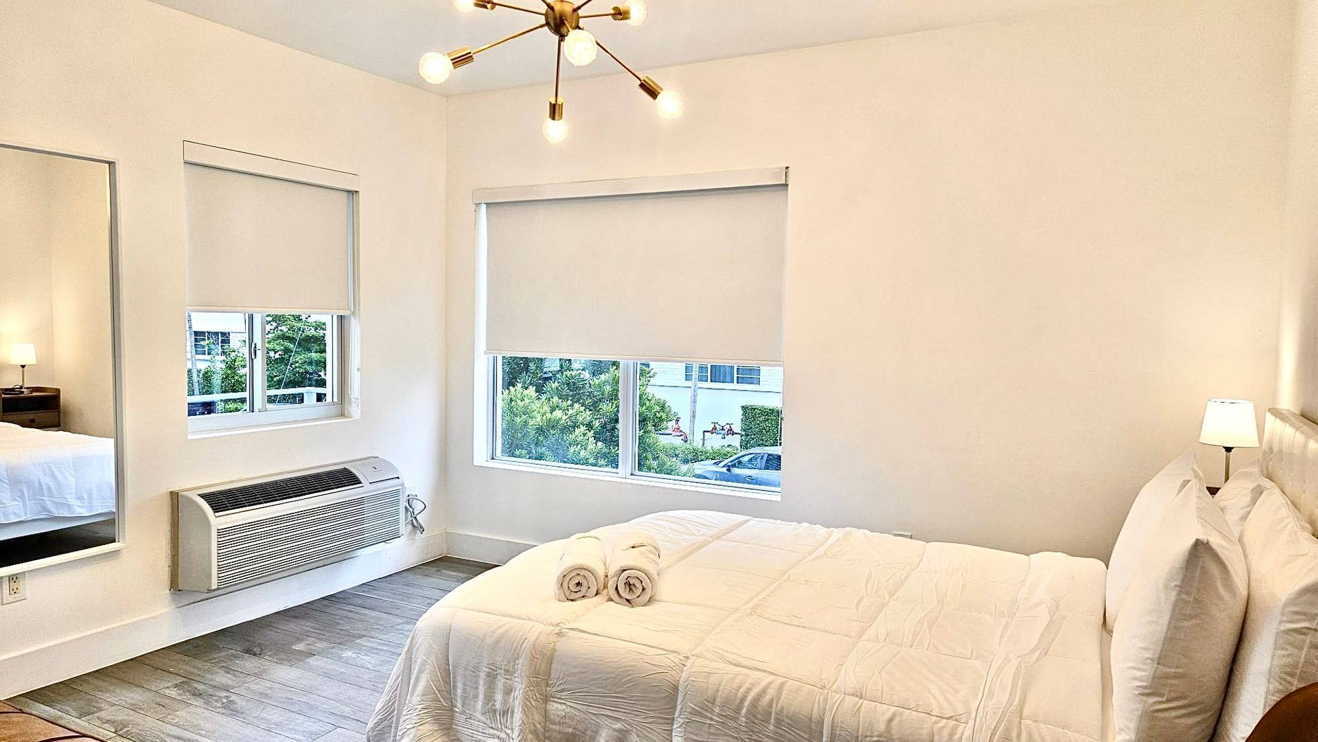 Bright bedroom with a bed, mirror, and window. The room is white with a modern light fixture.