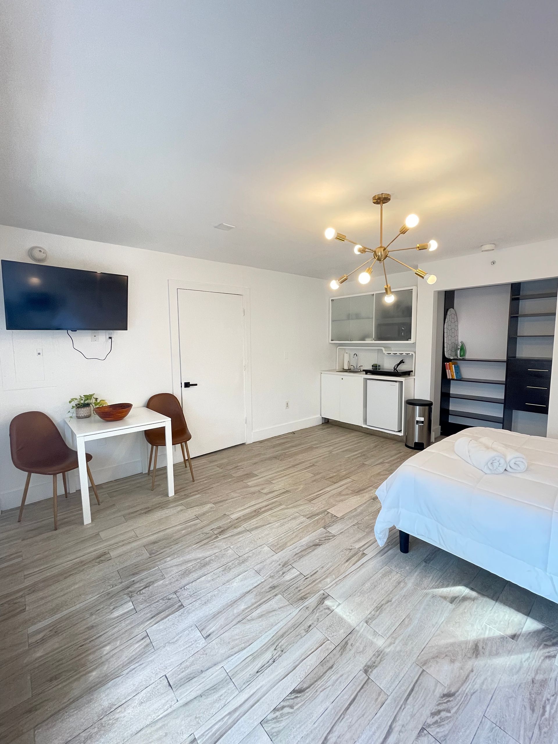 A studio apartment with a bed, small dining table, kitchenette, and a TV on the wall.