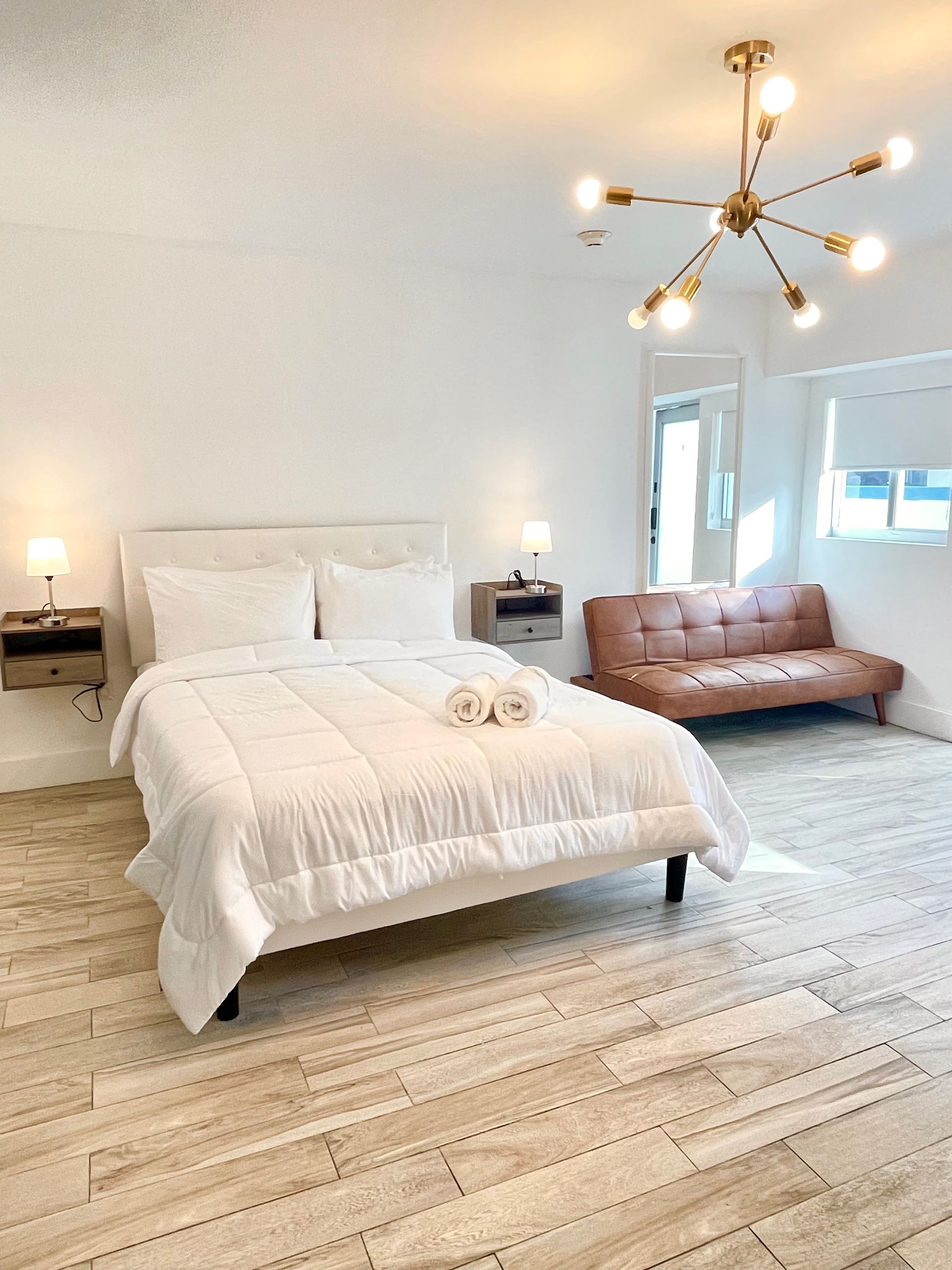 Bright, white bedroom with a bed, sofa, and modern light fixture. Neutral-toned wood floor.