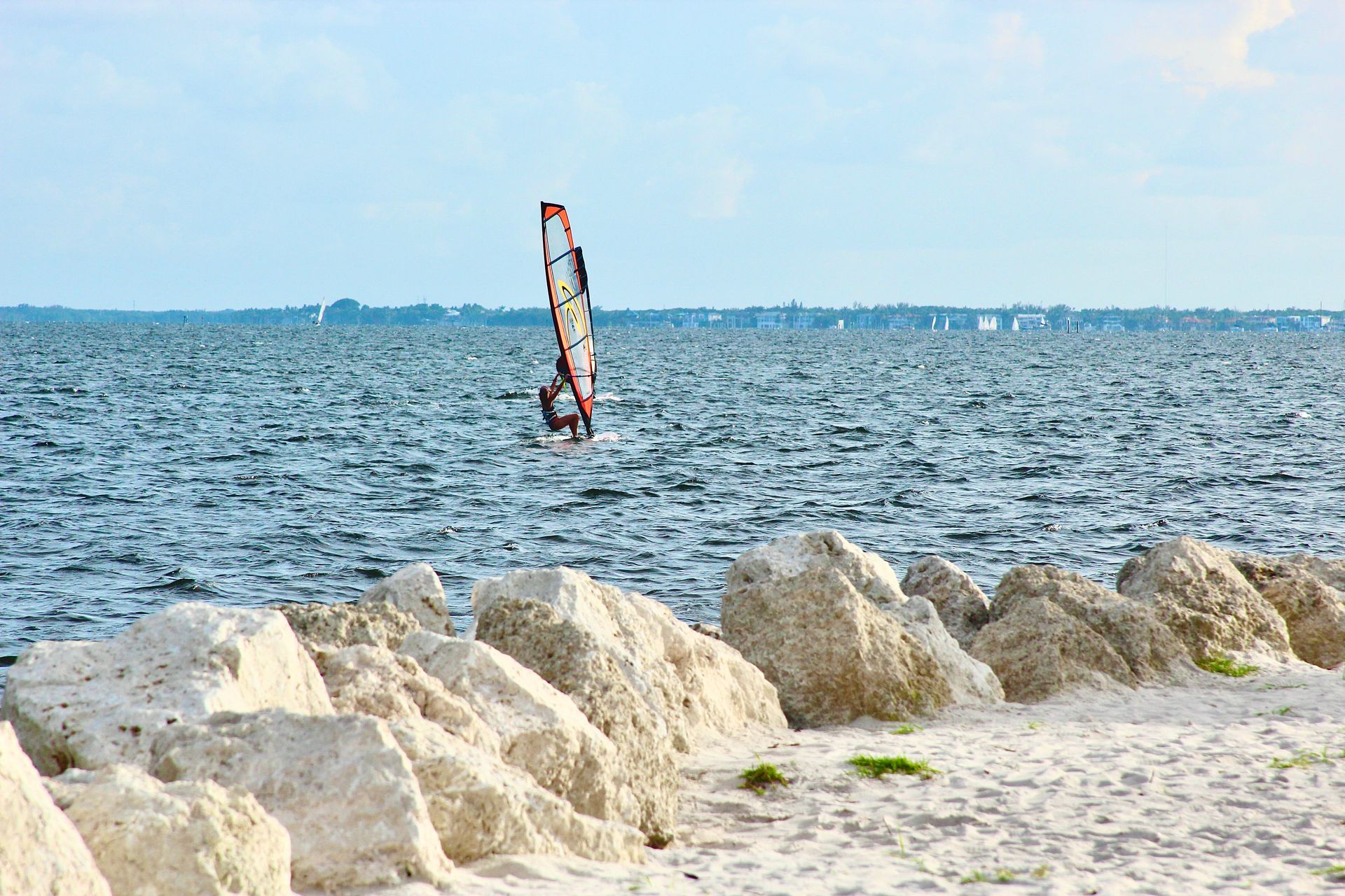 Windsurfer on blue water, near a rocky shore, under a light blue sky.