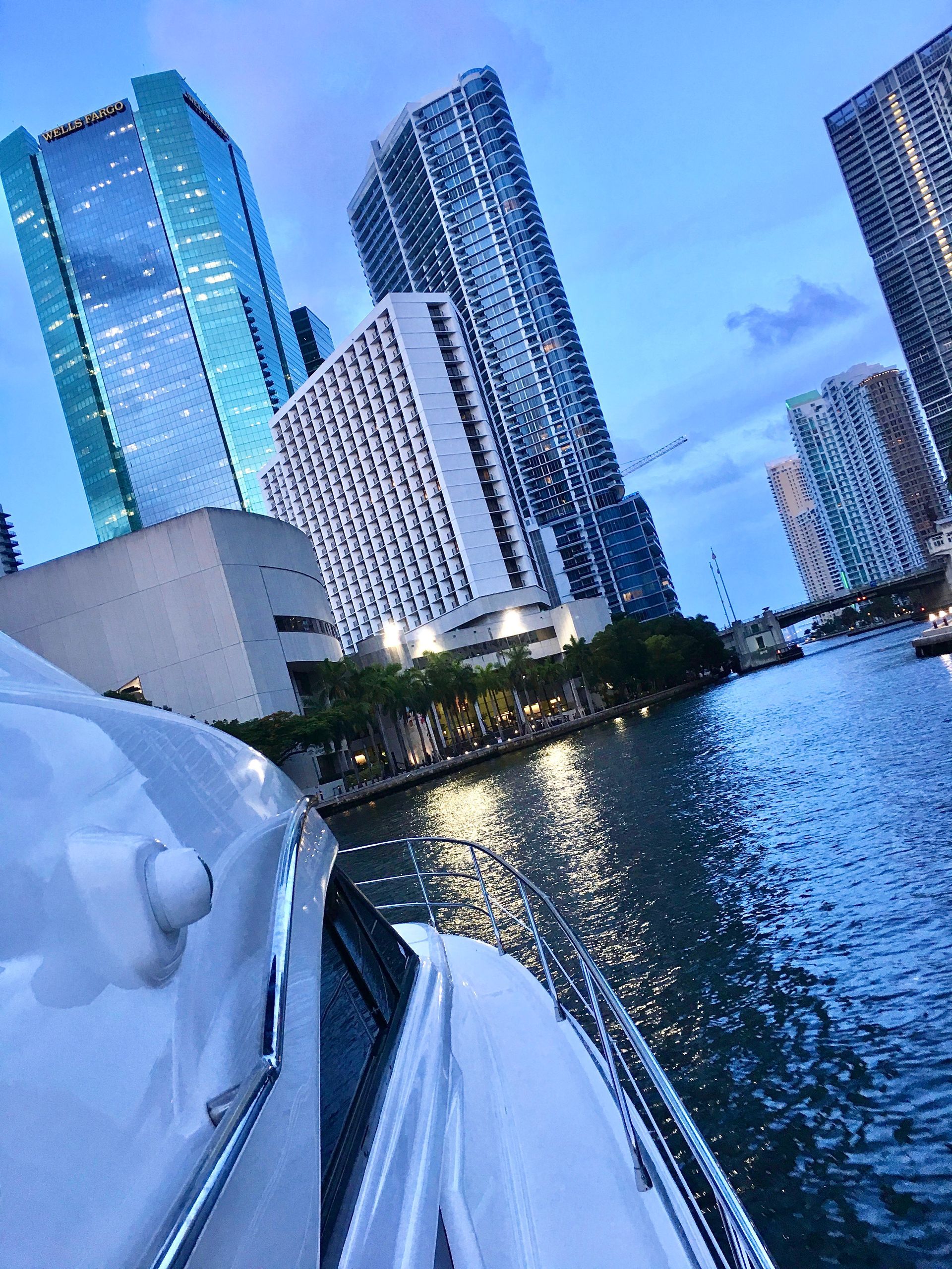 Boat cruising down a canal lined with illuminated skyscrapers at dusk in Miami.