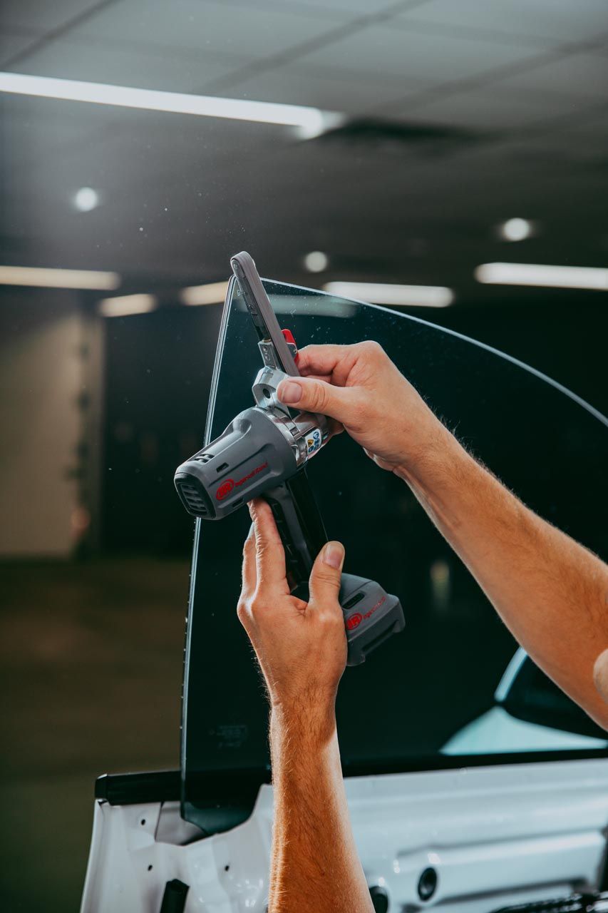 Person using a power tool on a tinted car window in a garage.