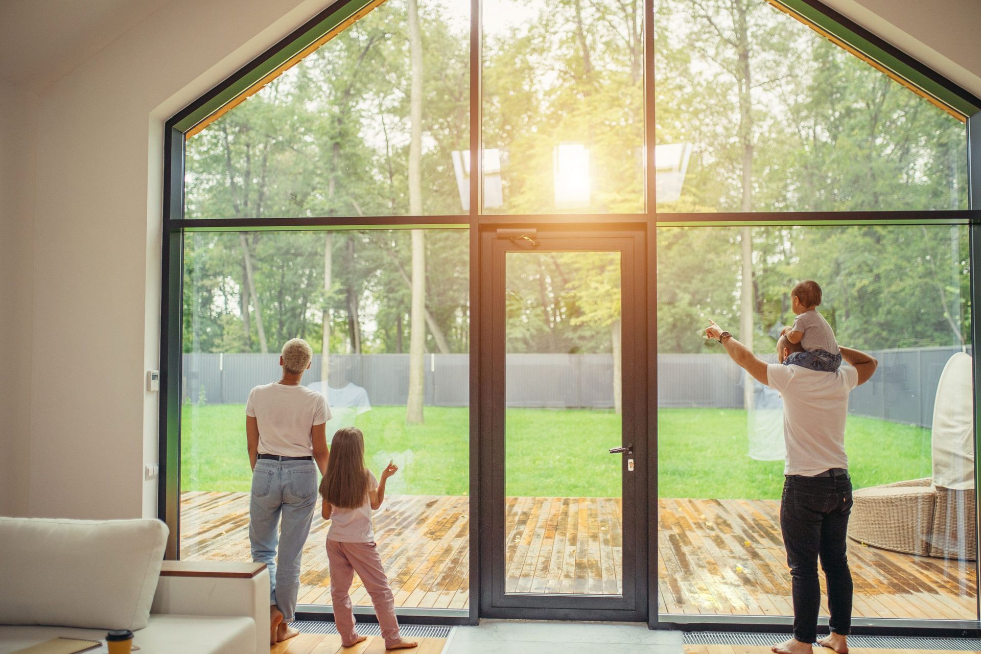 Family standing in a bright living room, looking out floor-to-ceiling windows at a sunny backyard.