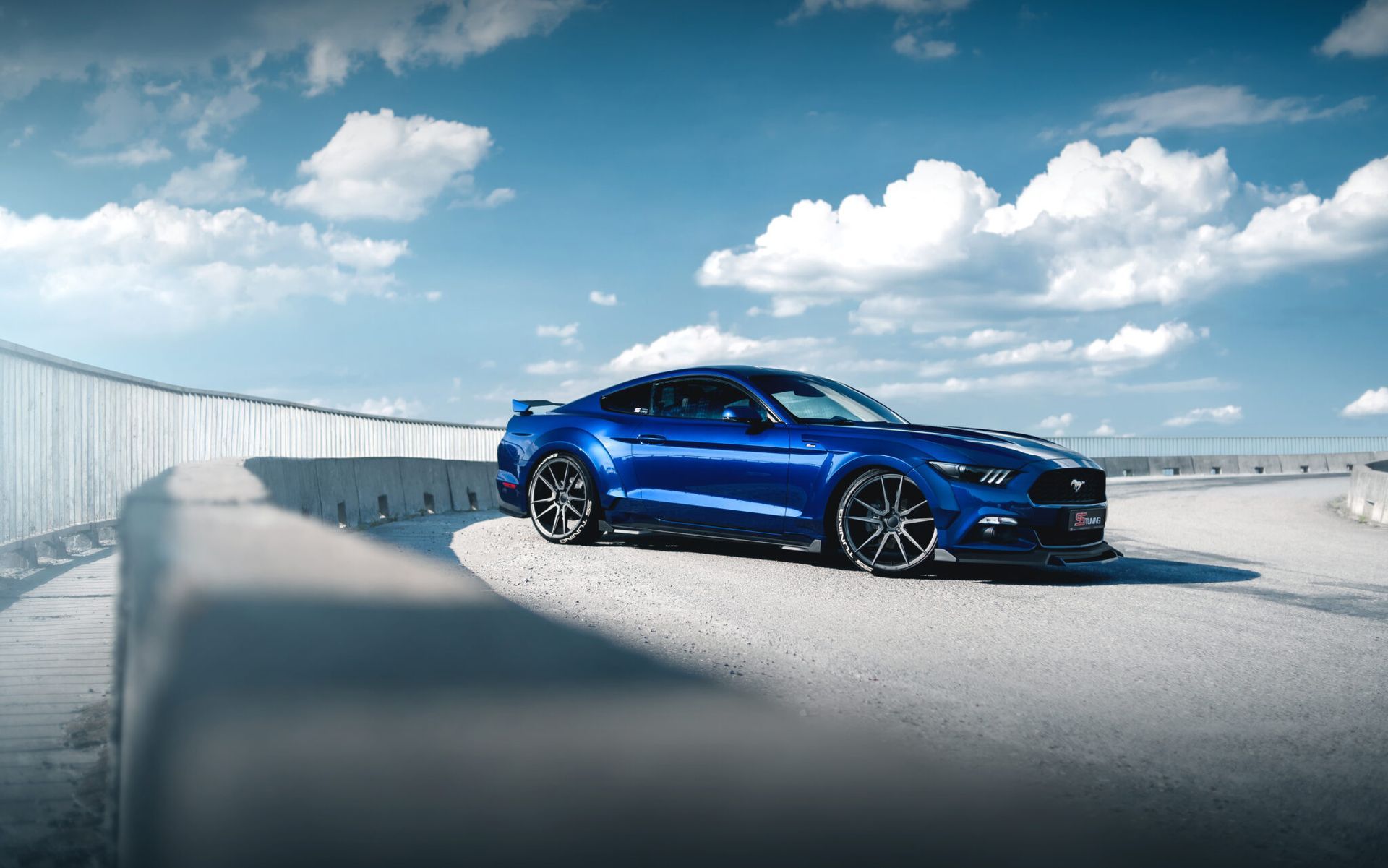 Blue sports sedan parked on a rooftop under a bright sky with clouds