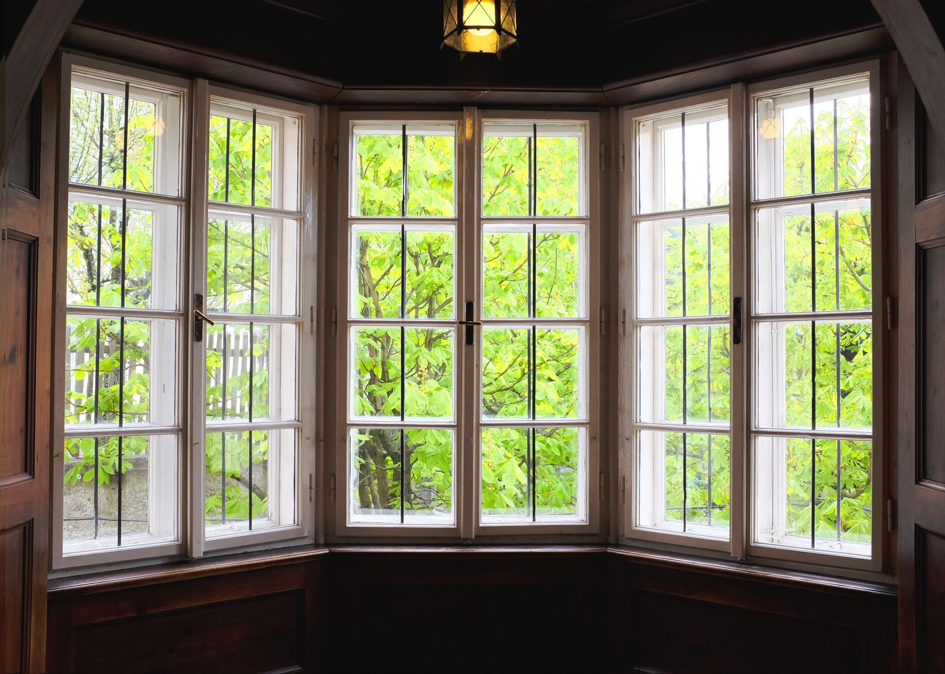 Wooden bay window with white panes overlooking green trees, lit by a hanging lantern