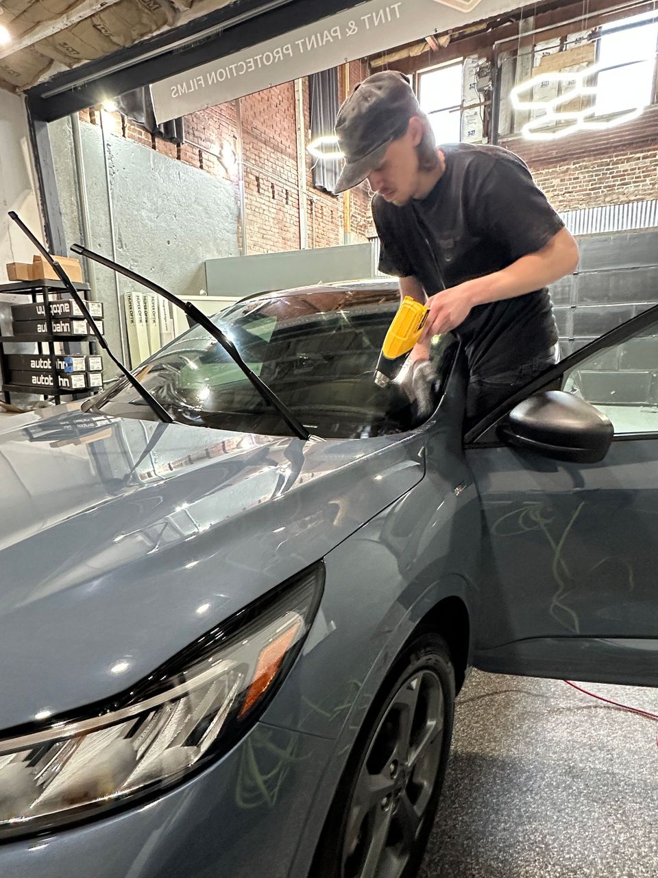 Person applying tint to a car window with a squeegee. The car is blue-gray. Indoor setting.