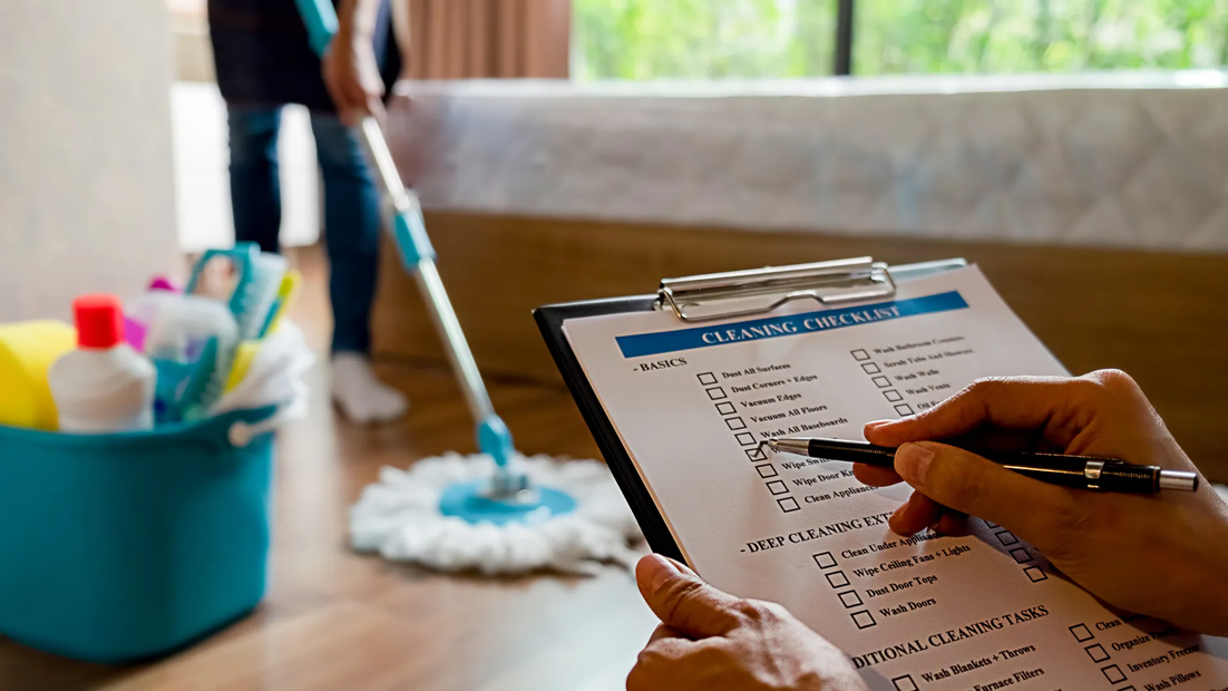 A person is writing on a clipboard in front of a mop and bucket of cleaning supplies.