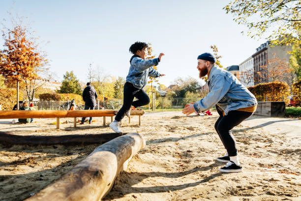 Child jumping over a log, with a man crouching to catch them in a sunny park setting.