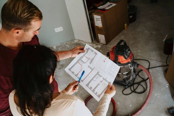 Couple reviewing blueprints in a room under renovation; man points with pen, woman observes.
