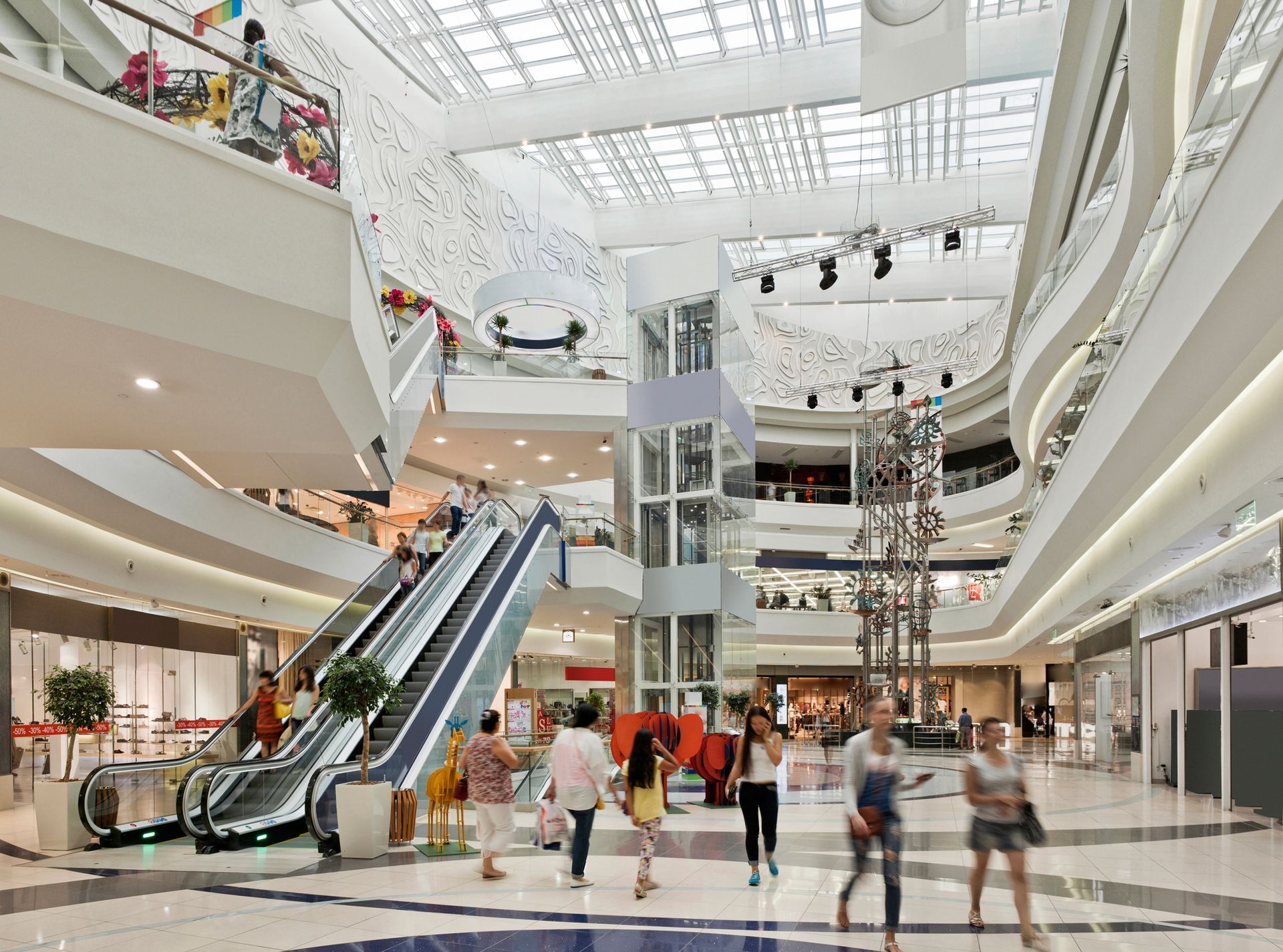 Interior view of a brightly lit shopping mall with escalators, glass elevators, and people walking around.