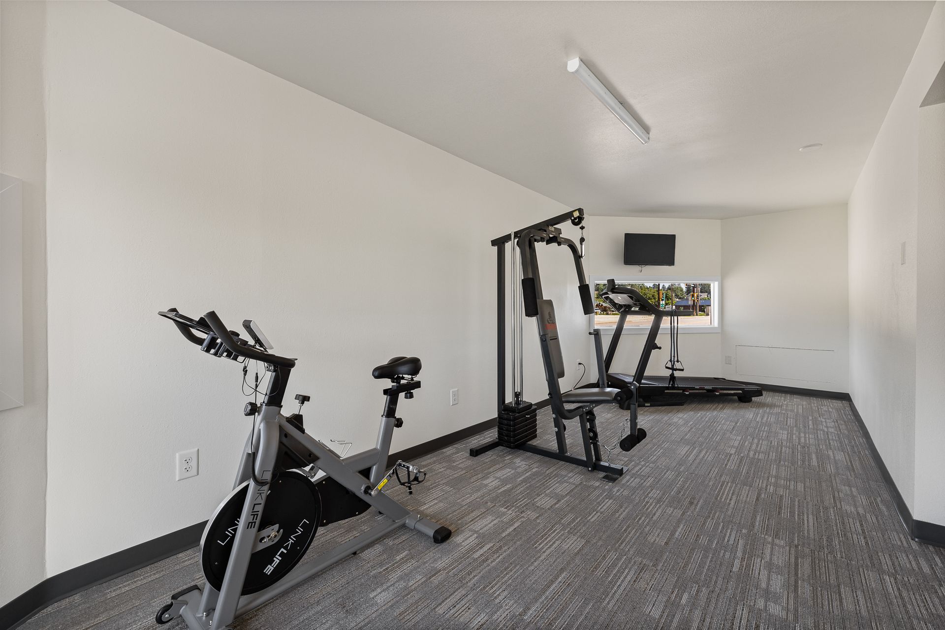 Exercise room with two stationary bikes and a weight machine on gray carpet.
