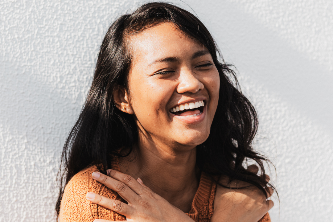 A woman is laughing with her eyes closed and her hand on her shoulder.