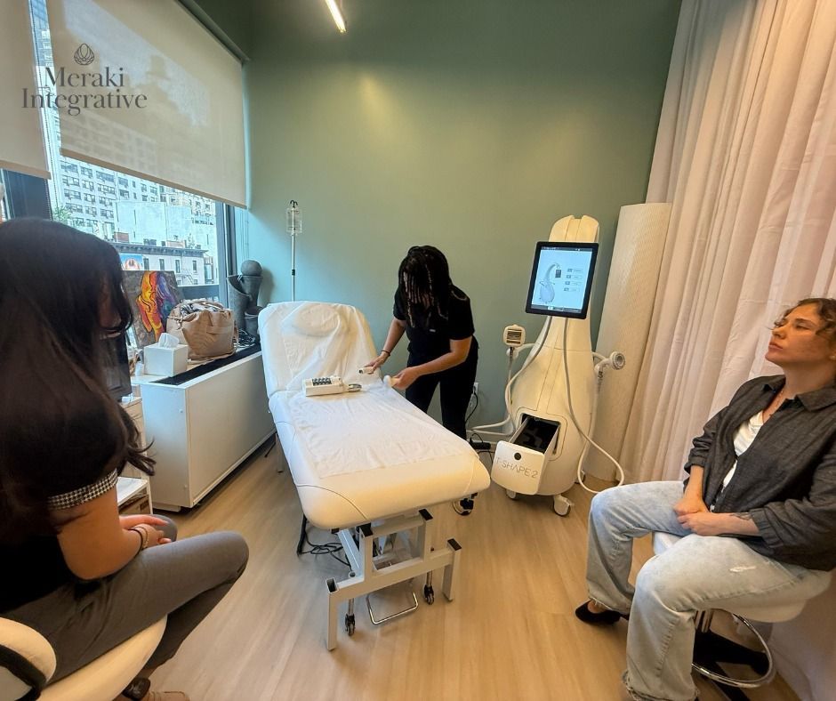 A woman prepares a treatment bed while another sits, in a modern medical office. A machine stands nearby.
