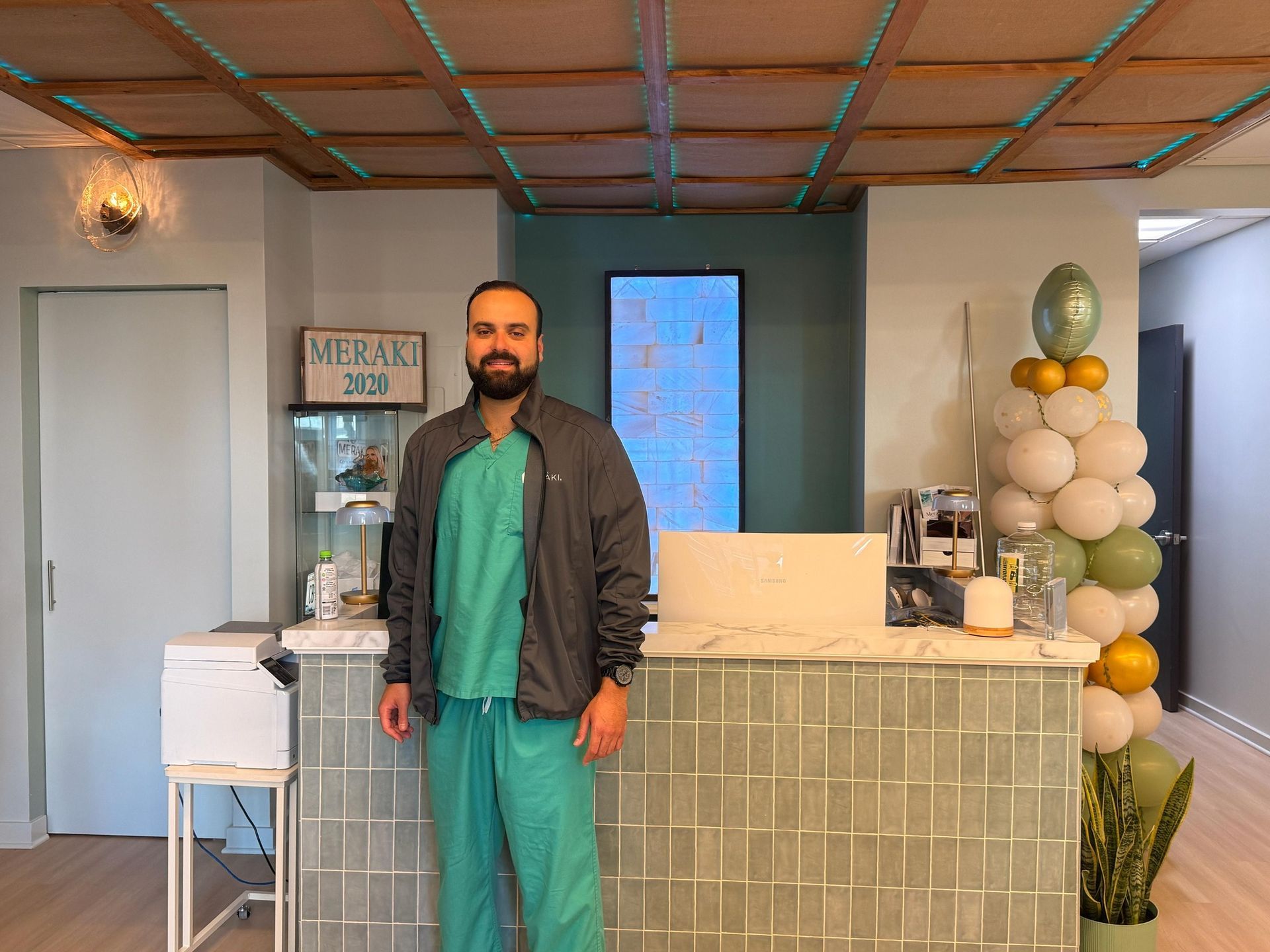 Man in scrubs stands at a reception desk in a light, modern office; green and gold balloon decor.