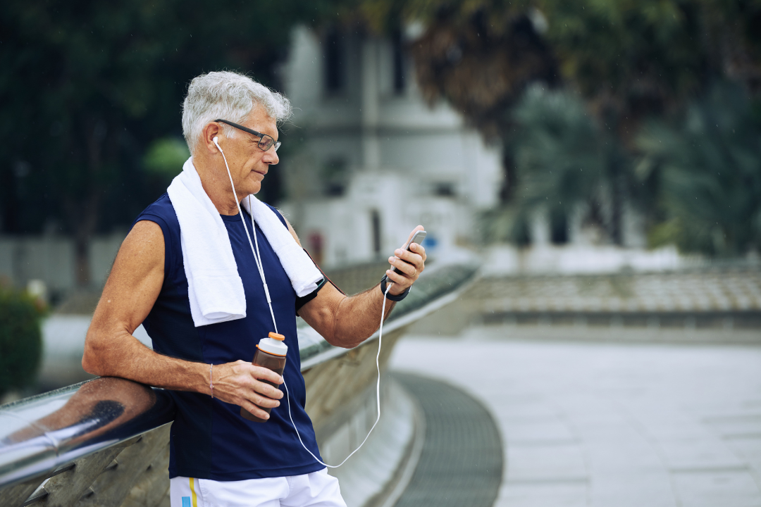 A person in athletic gear with a towel around their neck leans on a railing, using a phone with wired earbuds outdoors.