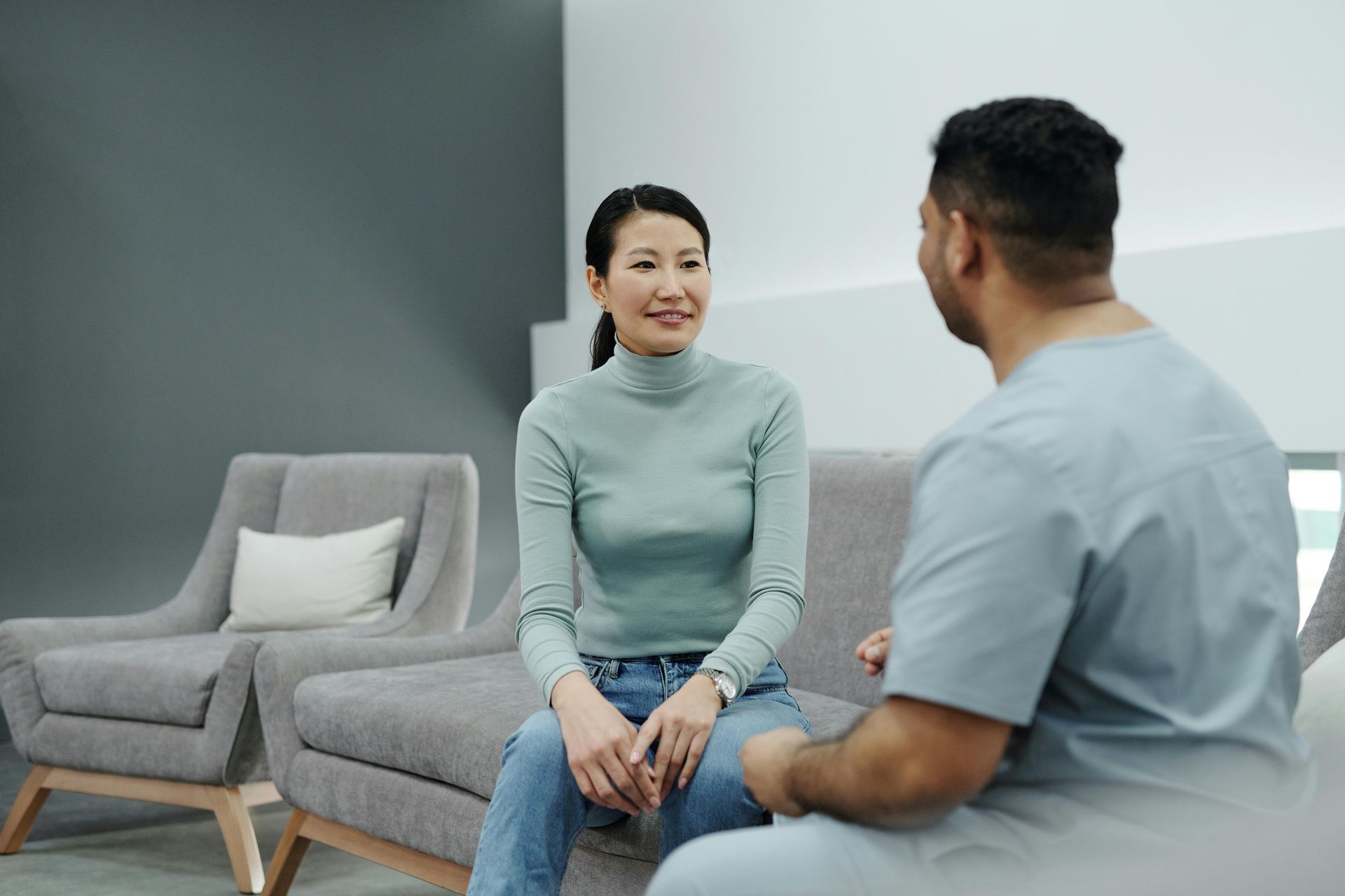 A person in a light blue mock-neck top sits in a grey armchair, speaking to a medical professional in scrubs.