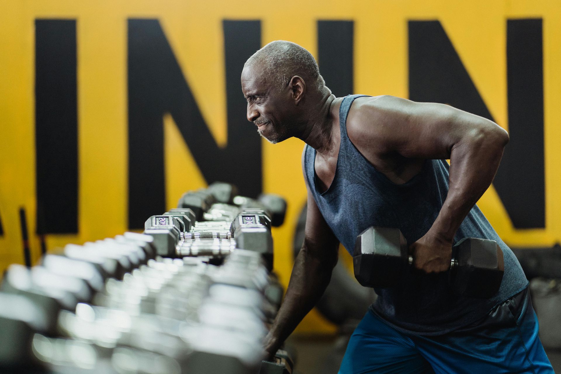 Man in gym lifting a dumbbell, sweating, working out. Row of dumbbells in front. Yellow wall behind.