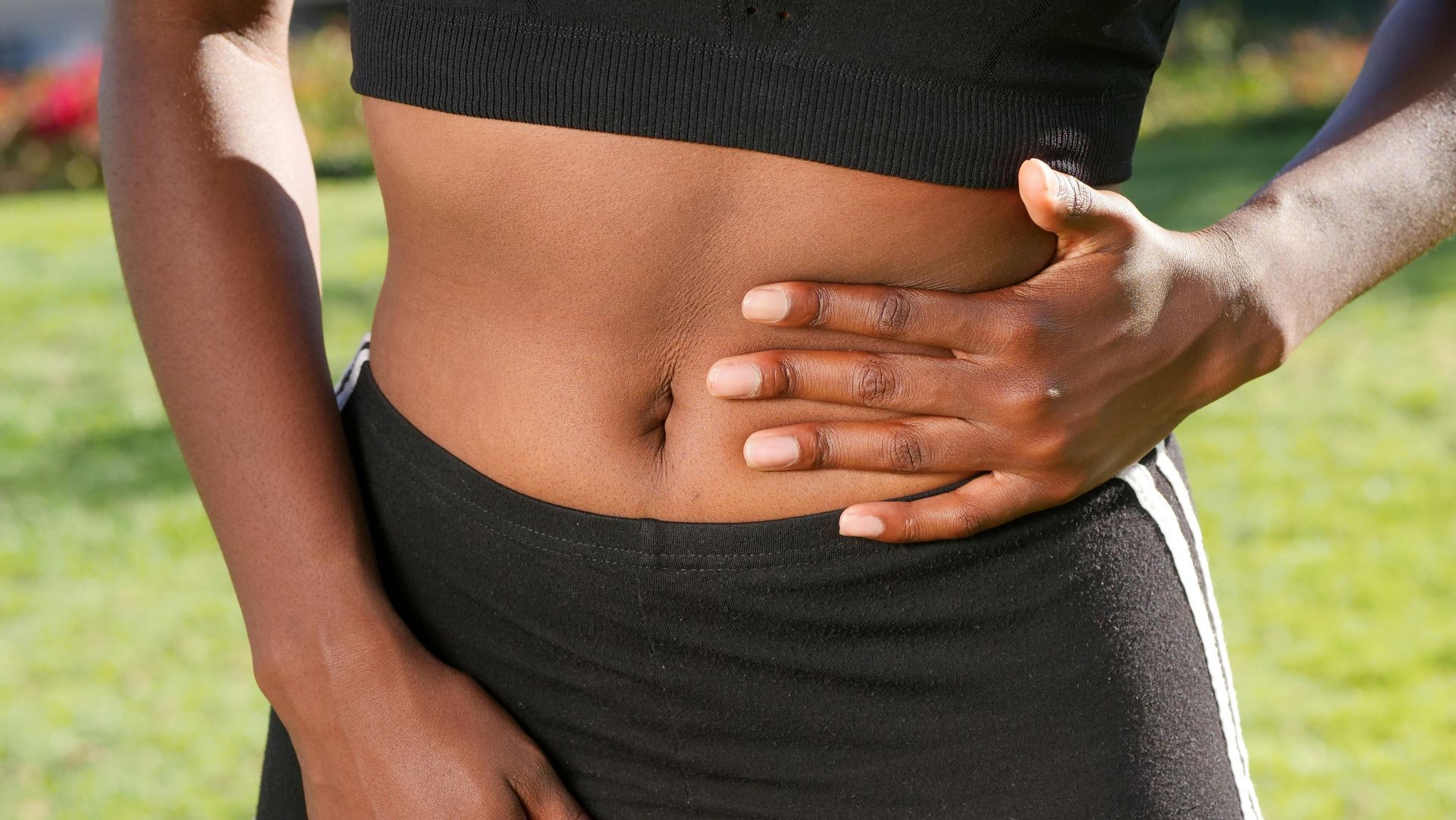 Close-up of a person holding their side with one hand, wearing a black top and pants outdoors on grass