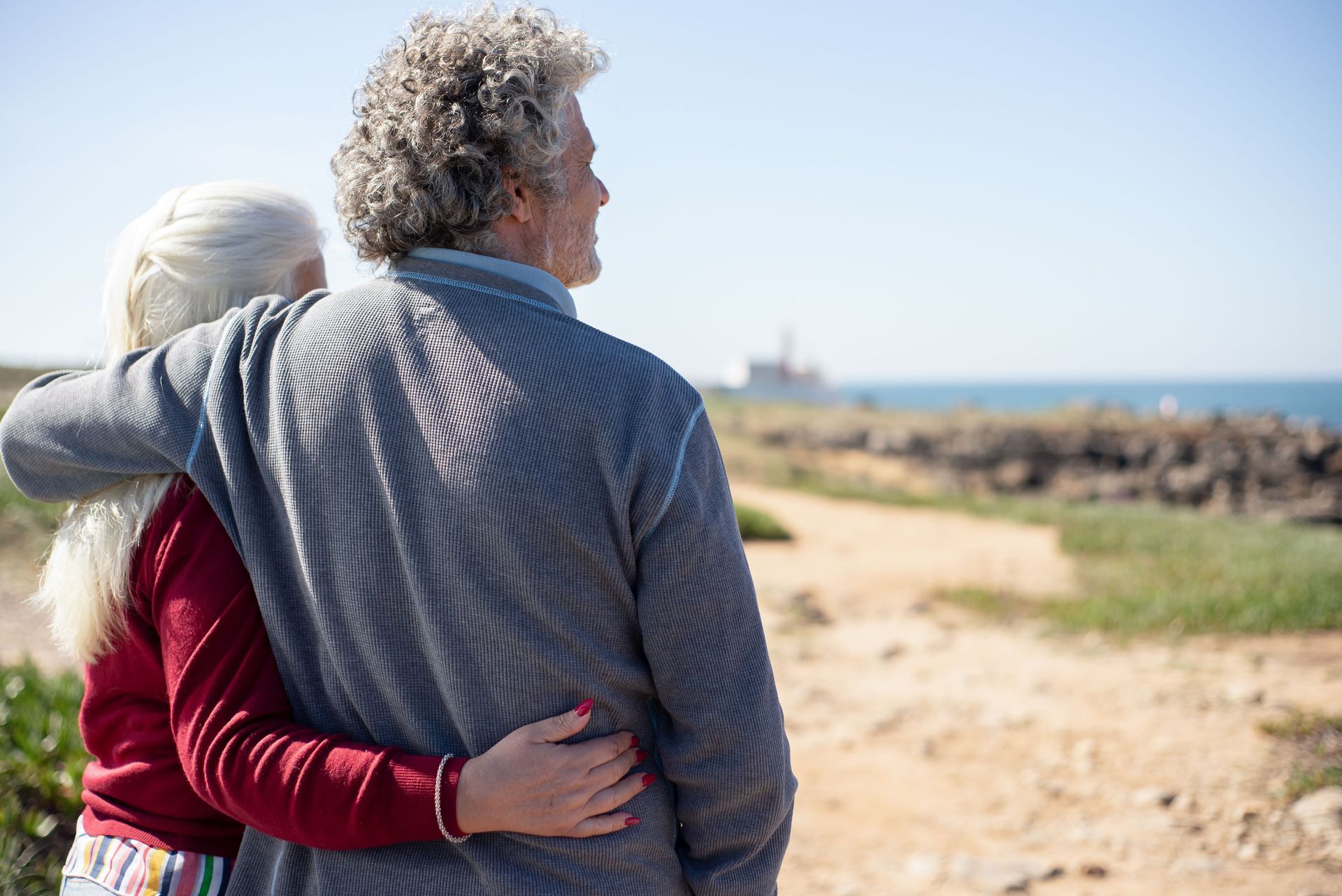 Two people embracing on a sandy coastal path beside open fields under a clear sky
