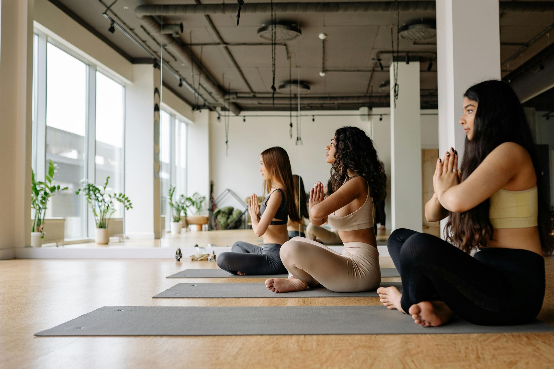 Three people seated on yoga mats in a bright studio, meditating beside large windows.