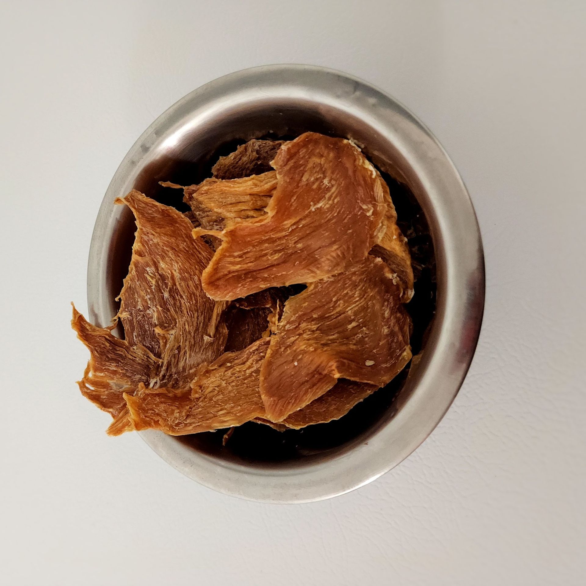 Dried meat pieces in a metal bowl on a white surface.