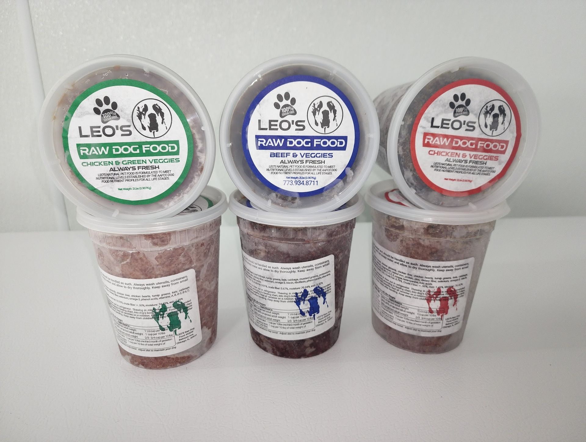 Containers of Leo's Raw Dog Food: green, blue, and red lids, stacked on a white surface.