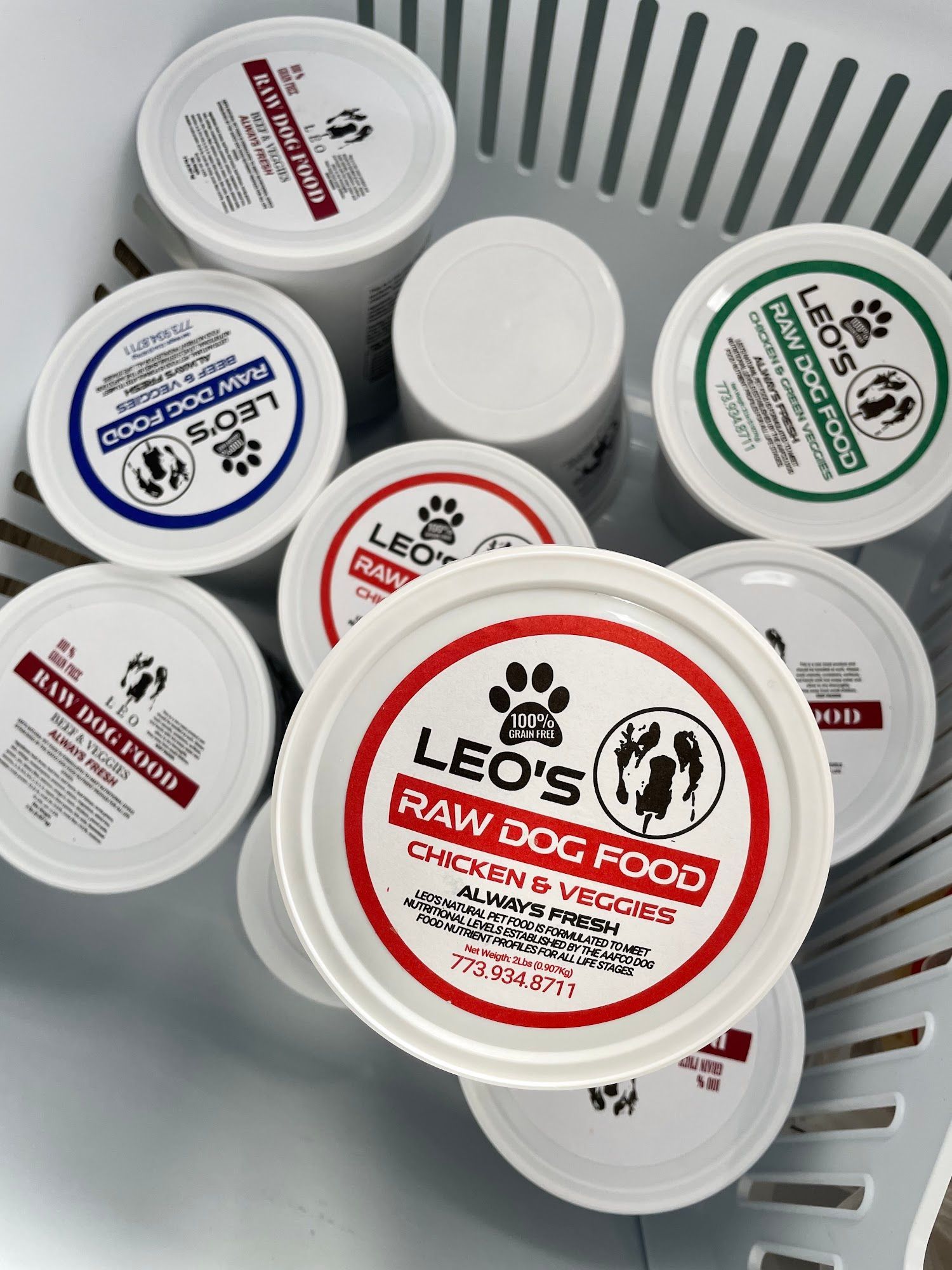 A basket filled with various containers of Leo's Raw Dog Food. The lids are white with colored labels.