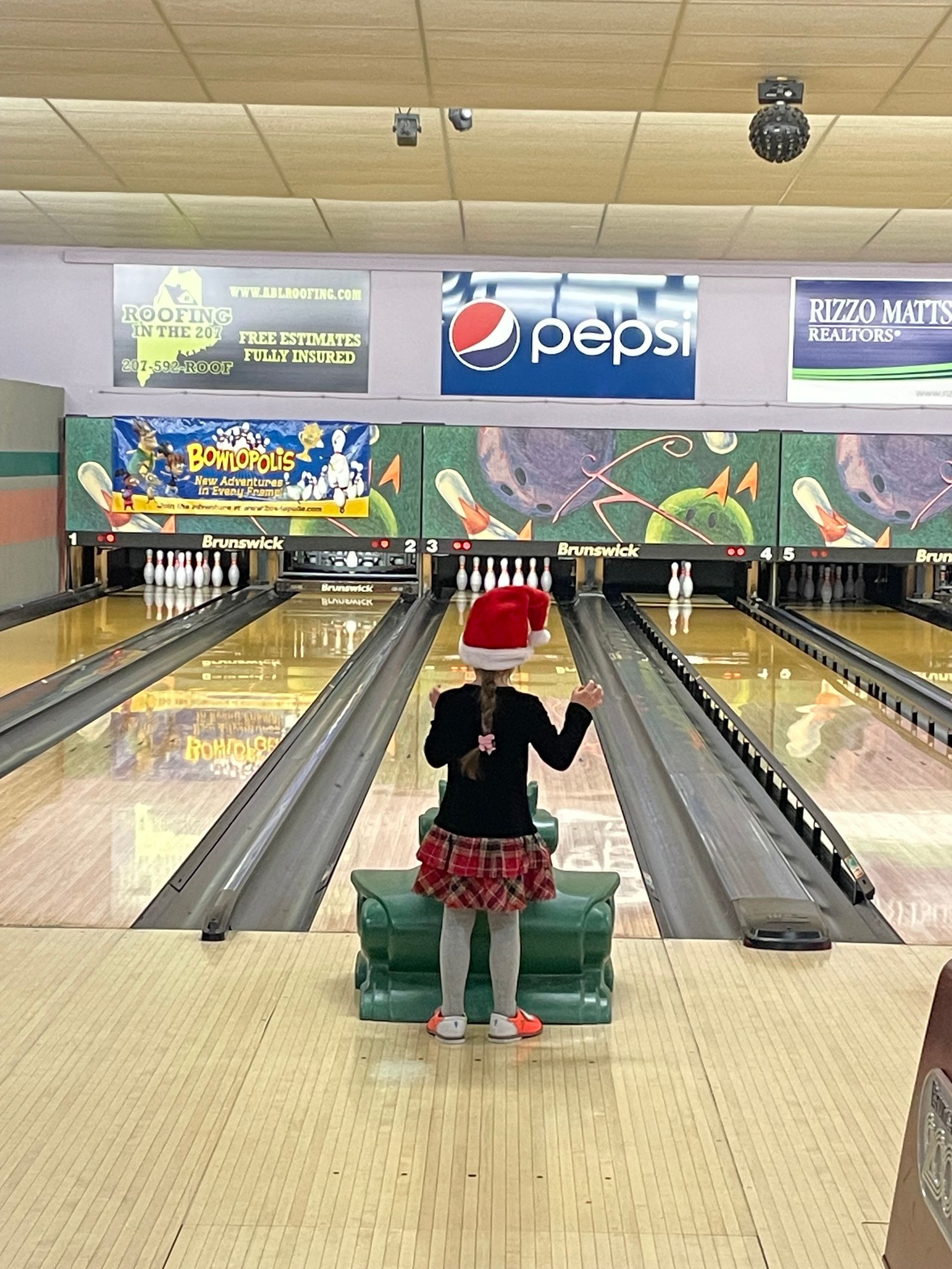 A little girl wearing a santa hat is standing in a bowling alley.