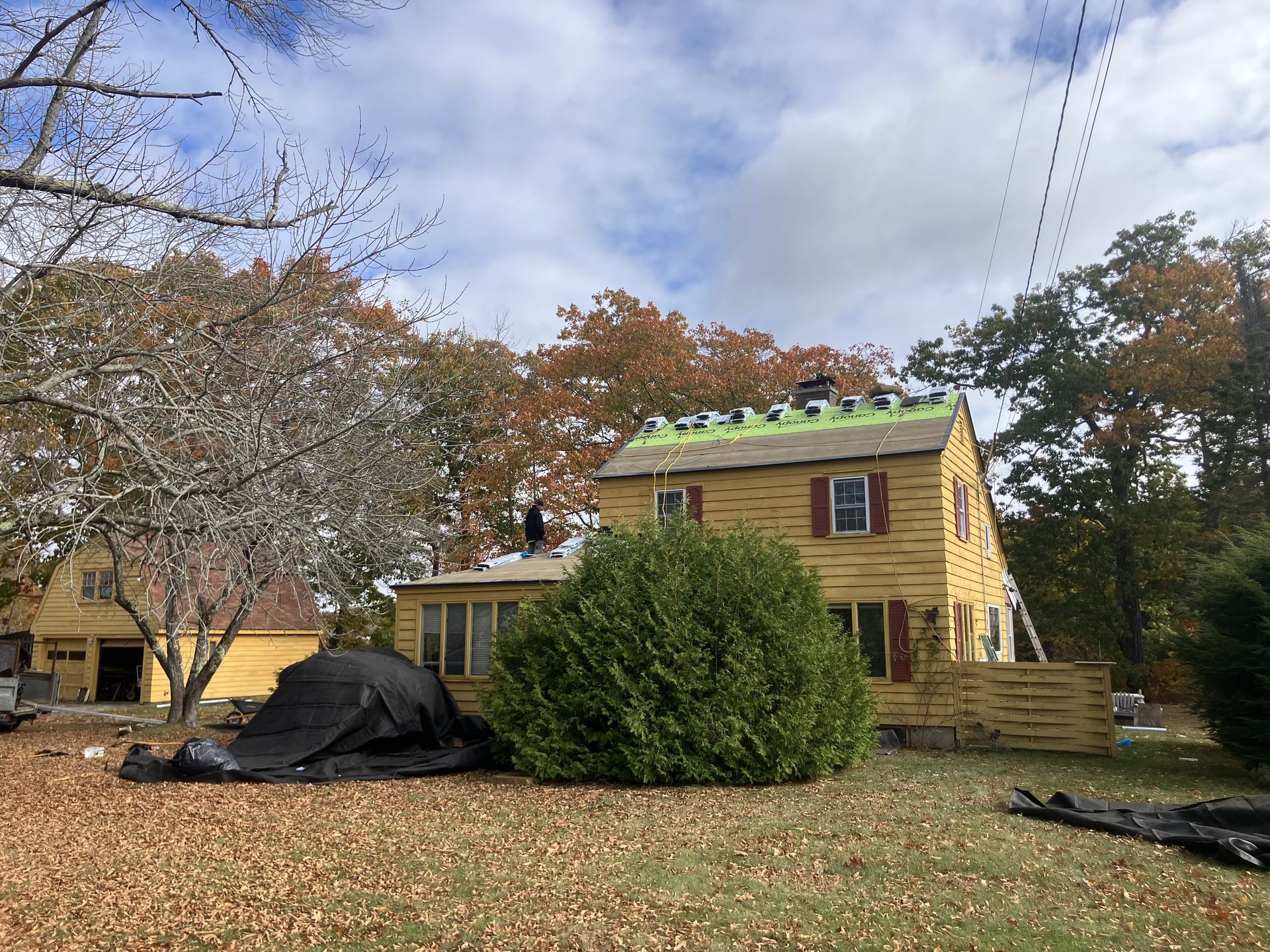 House with partially replaced roof under cloudy sky, surrounded by fall foliage. A person is on the roof.