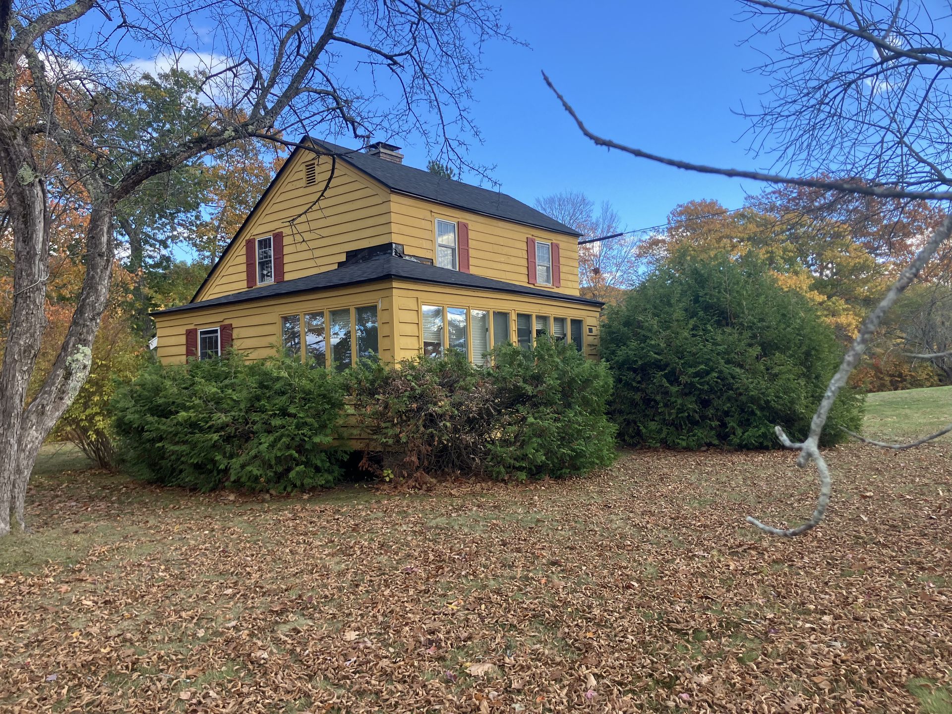 Yellow house with dark roof and red shutters, surrounded by fall foliage.