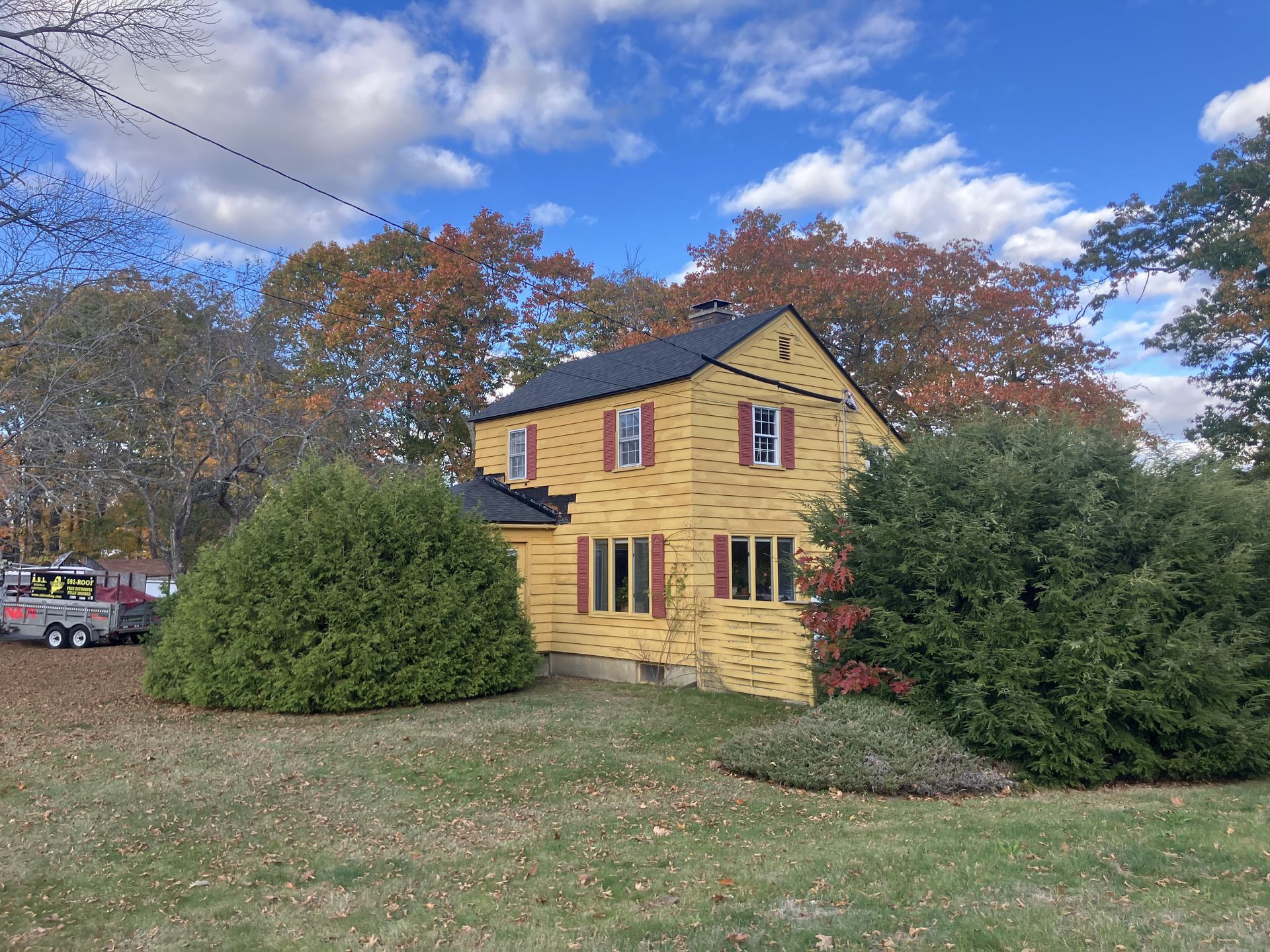 Yellow house with red shutters, nestled among trees with fall foliage, under a blue sky with clouds.