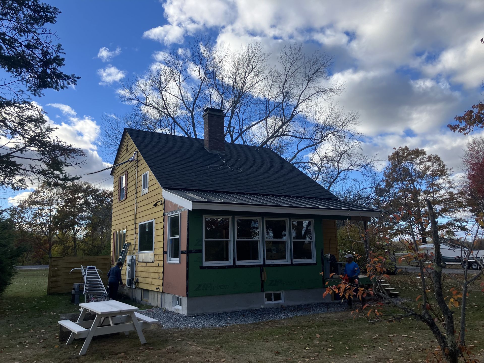 House under construction, part yellow, part green, black roof, small porch, cloudy sky.